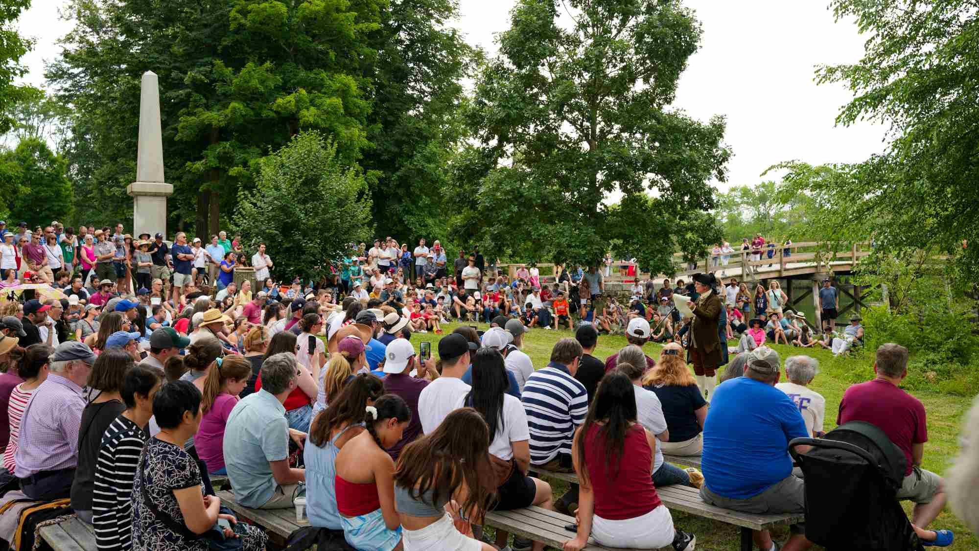 Large crowd seated outdoors at a historical park with an obelisk and wooden bridge in the background.