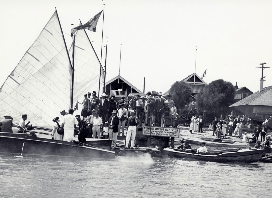 Black-and-white historical photo of a crowd on a dock beside a sailboat with boathouses in the background.