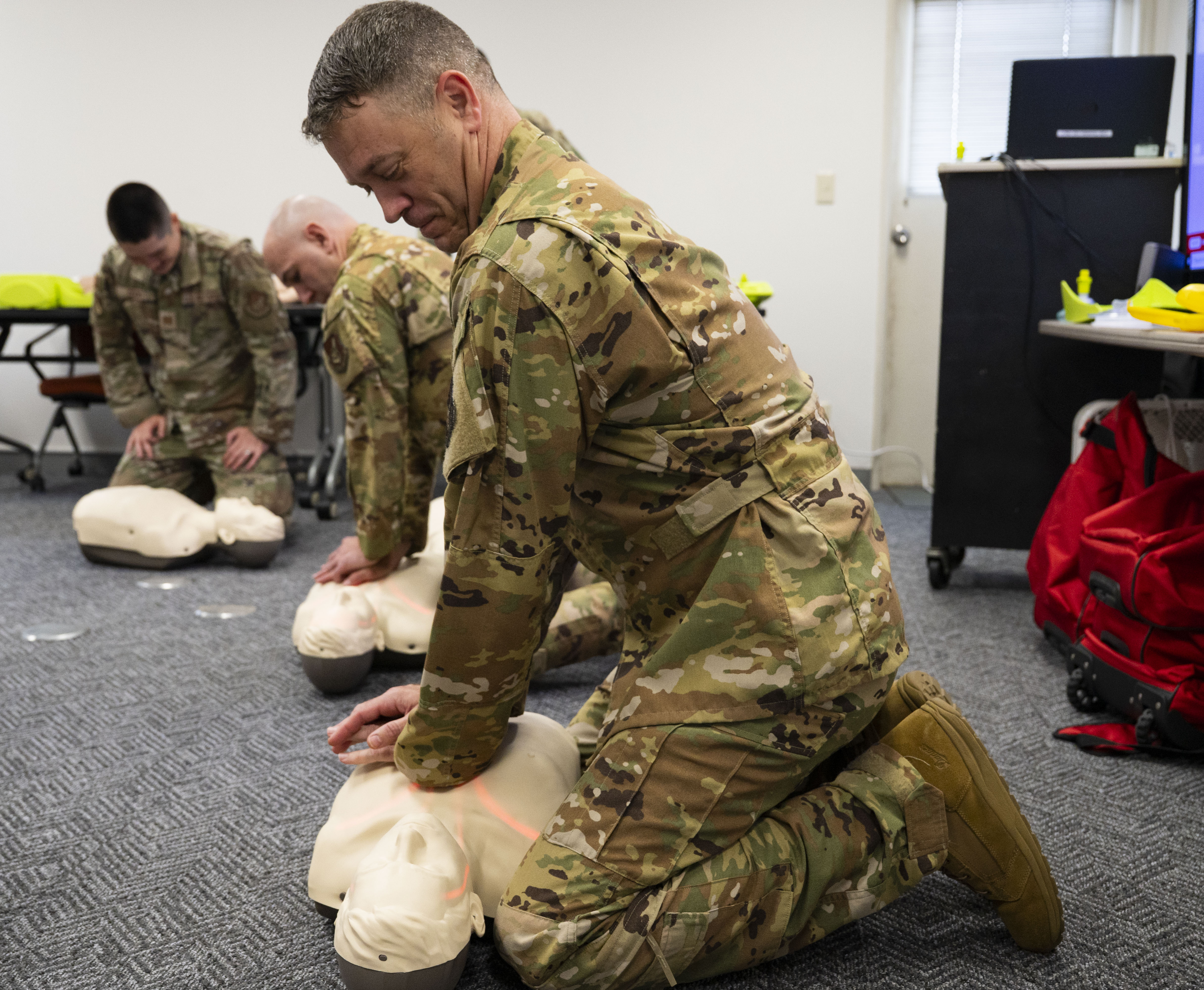 Person in military uniform kneeling and performing chest compressions on a CPR training mannequin.