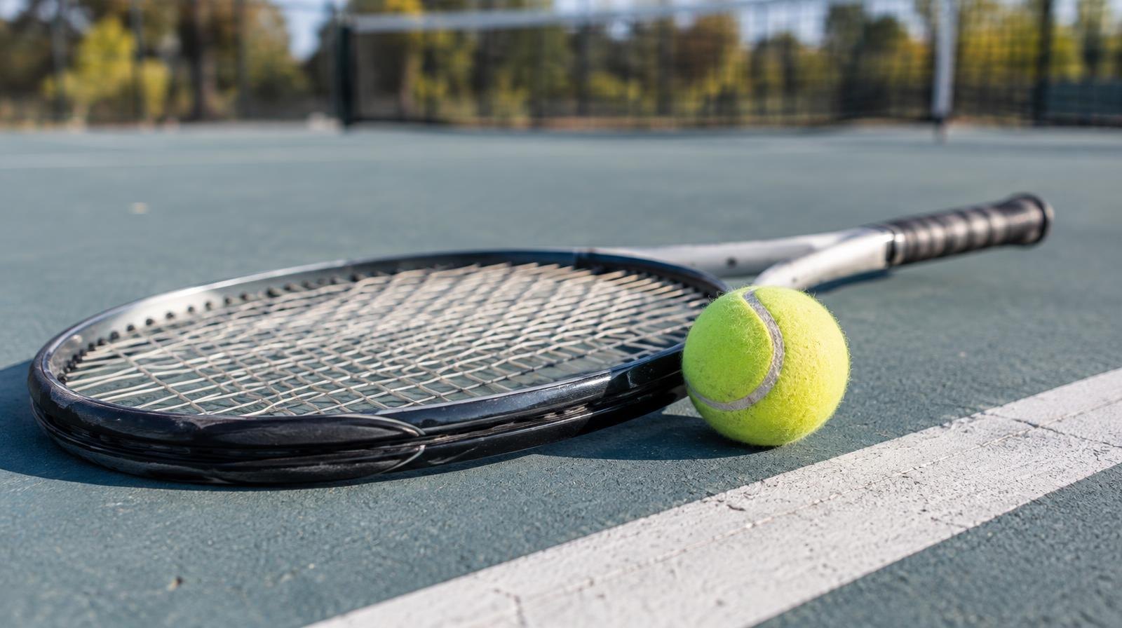 A tennis racket resting on a hard court beside a yellow tennis ball near the baseline.