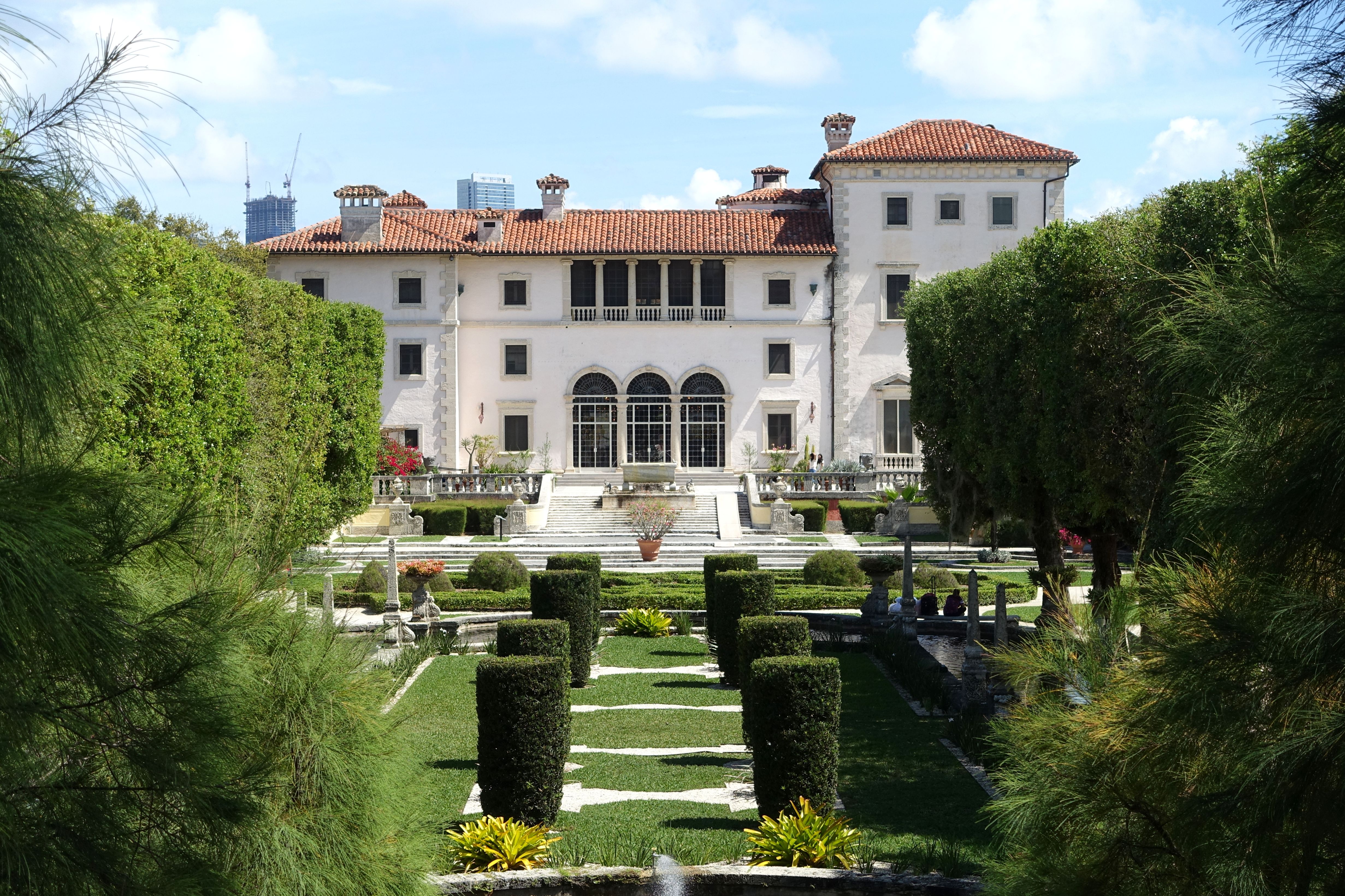 White Italian Renaissance villa with formal hedged gardens and city skyline behind.