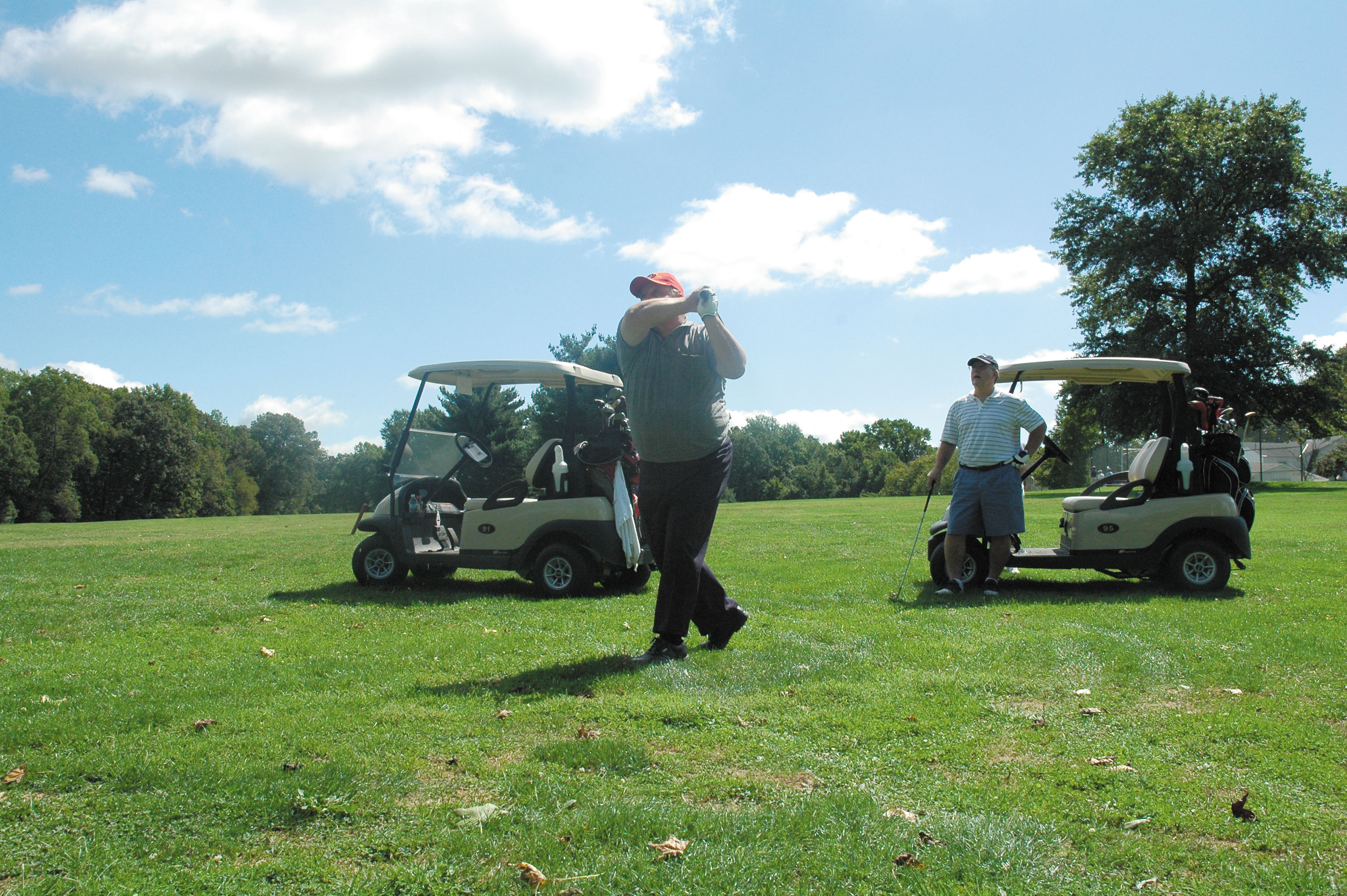 Two golfers on a sunny course, one mid-swing, with golf carts nearby.