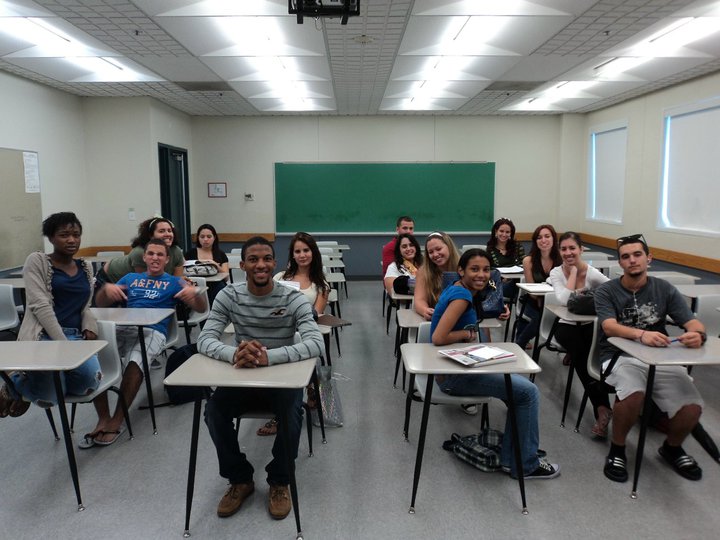 Students seated at desks in a classroom with a green chalkboard and blue walls.