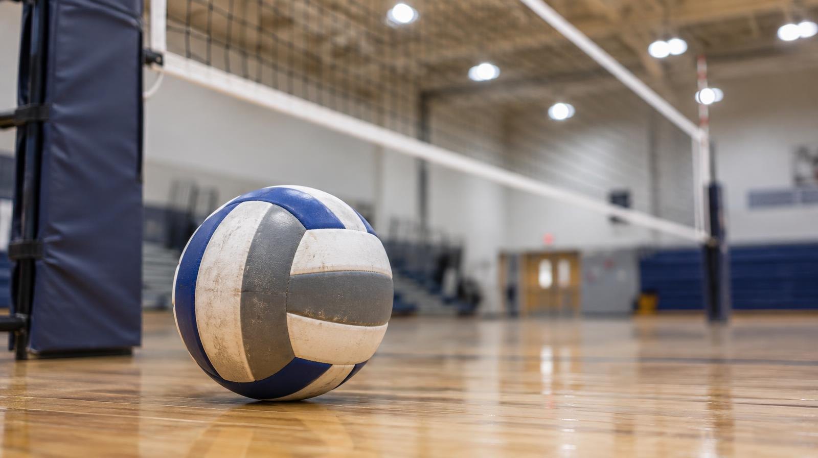 A volleyball resting on a hardwood gym floor beside a net post.