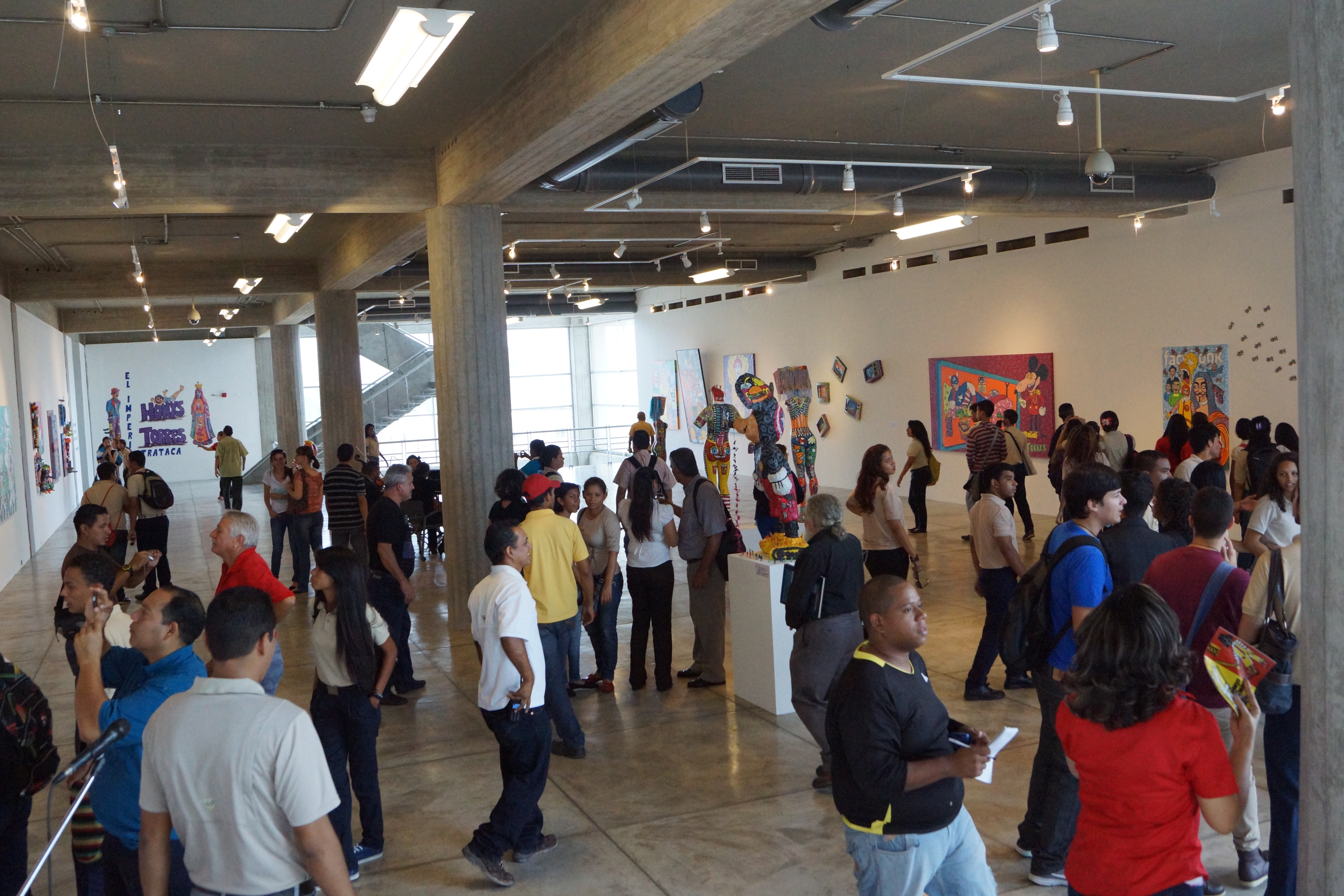 Crowds of visitors walk through a contemporary art gallery viewing colorful paintings.