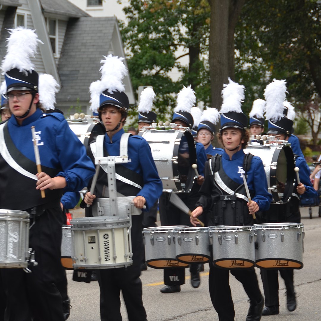 Marching band members in blue uniforms with white shakos playing snare drums on a street.