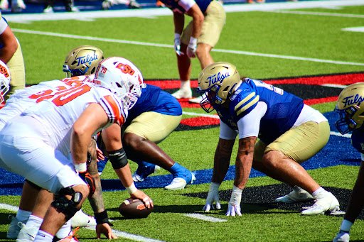 Football players in gold and blue uniforms face off against white and orange opponents on blue turf.