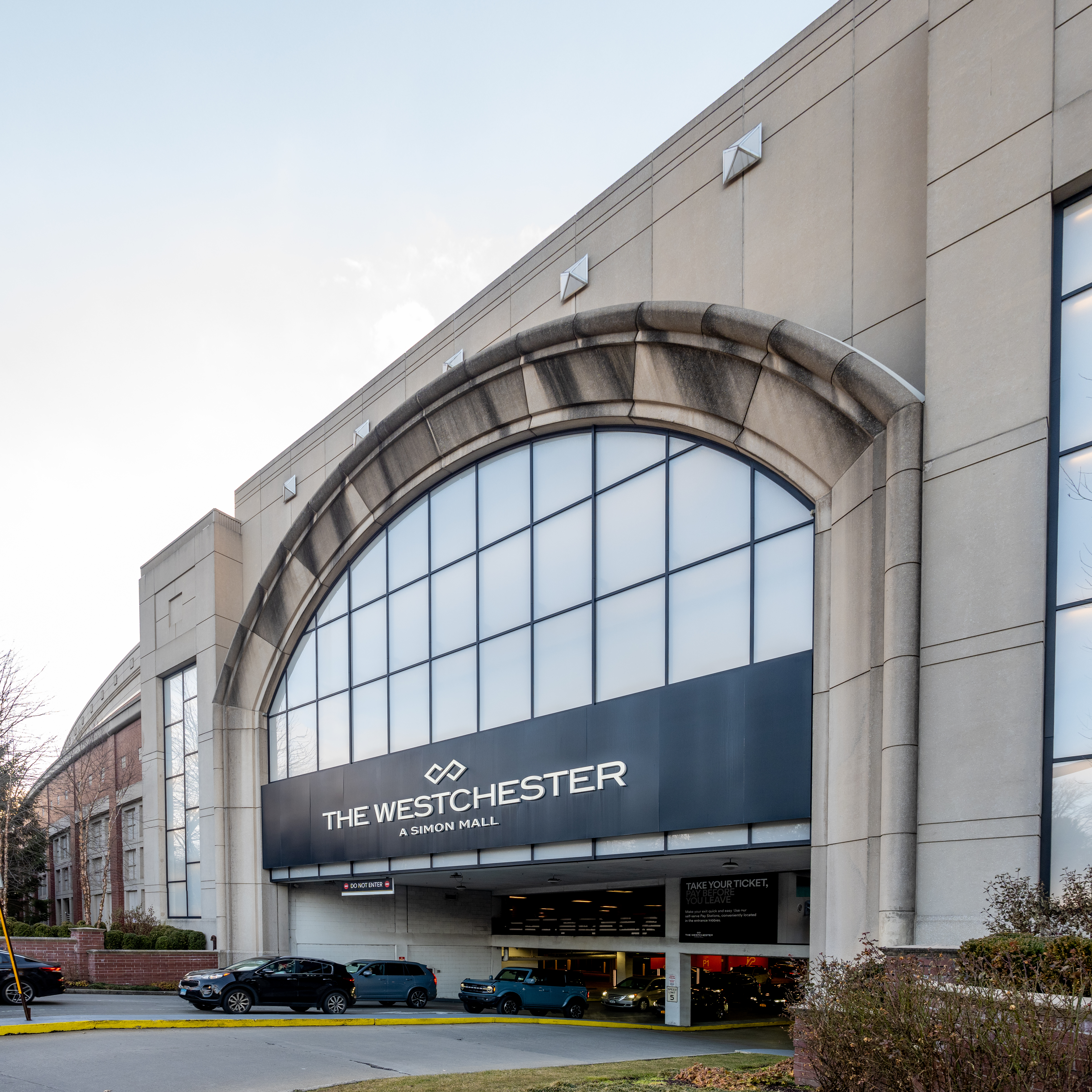 Exterior of The Westchester mall showing a large arched window entrance and parked cars.