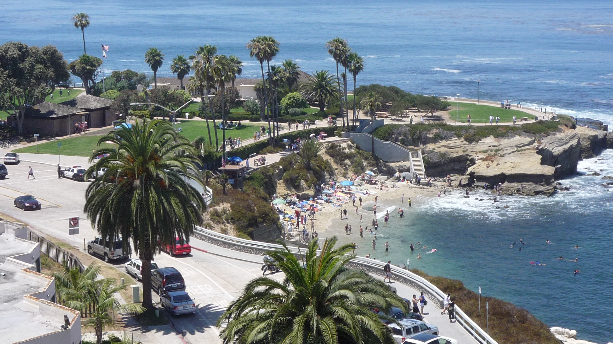 Aerial view of a coastal cove with sandy beach, palm trees, and a grassy park.