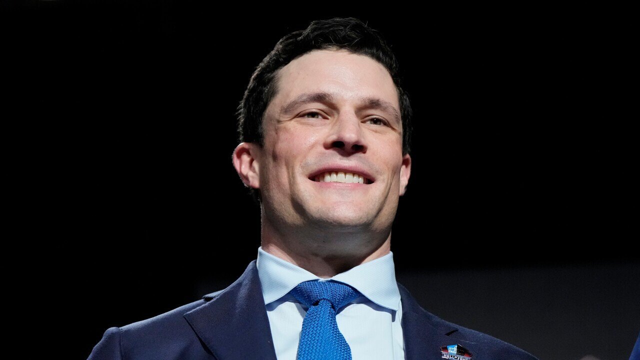 Smiling man in a dark navy suit and light blue tie against a dark background.