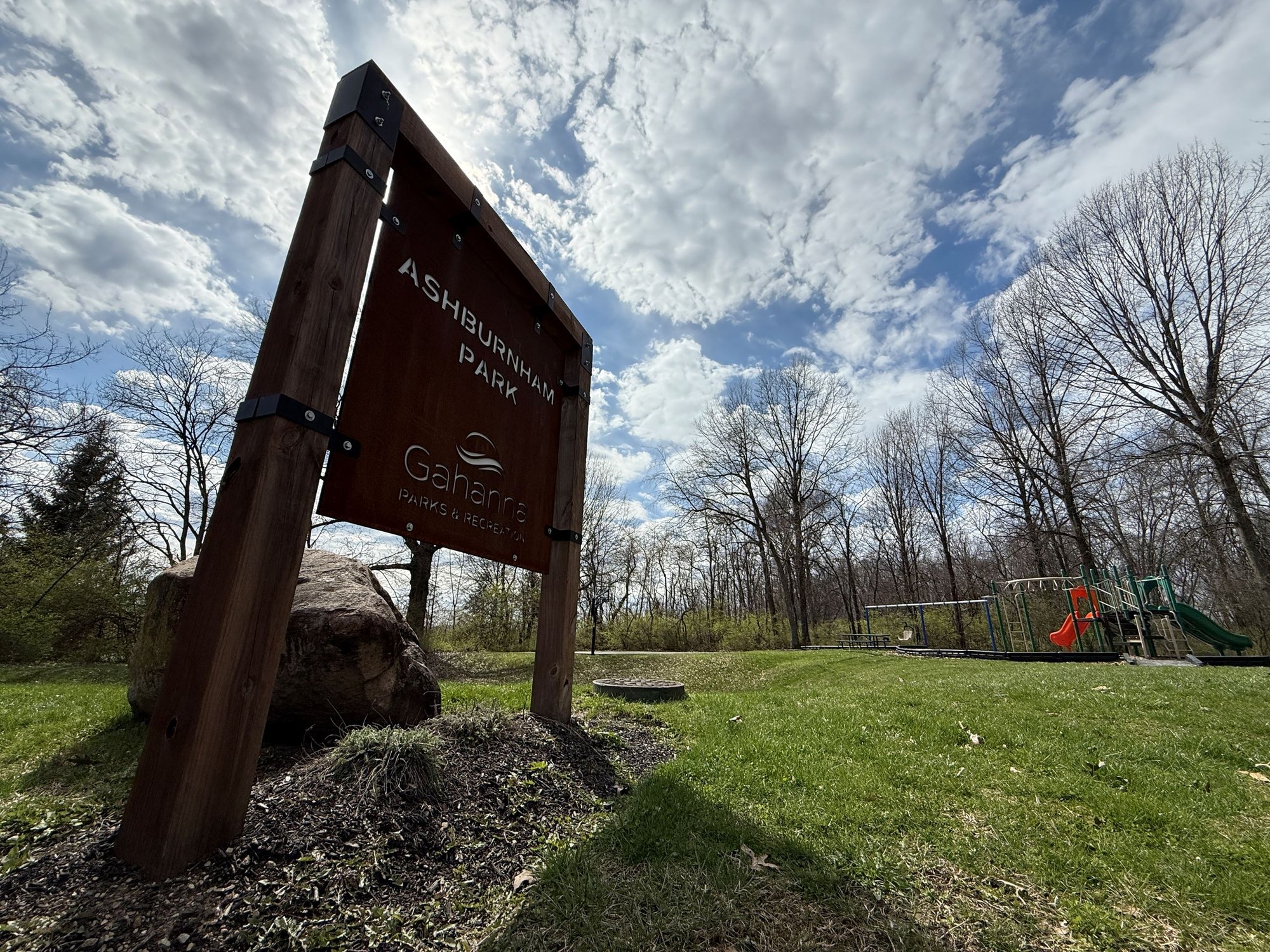 Wooden park entrance sign with a playground and trees visible behind it.