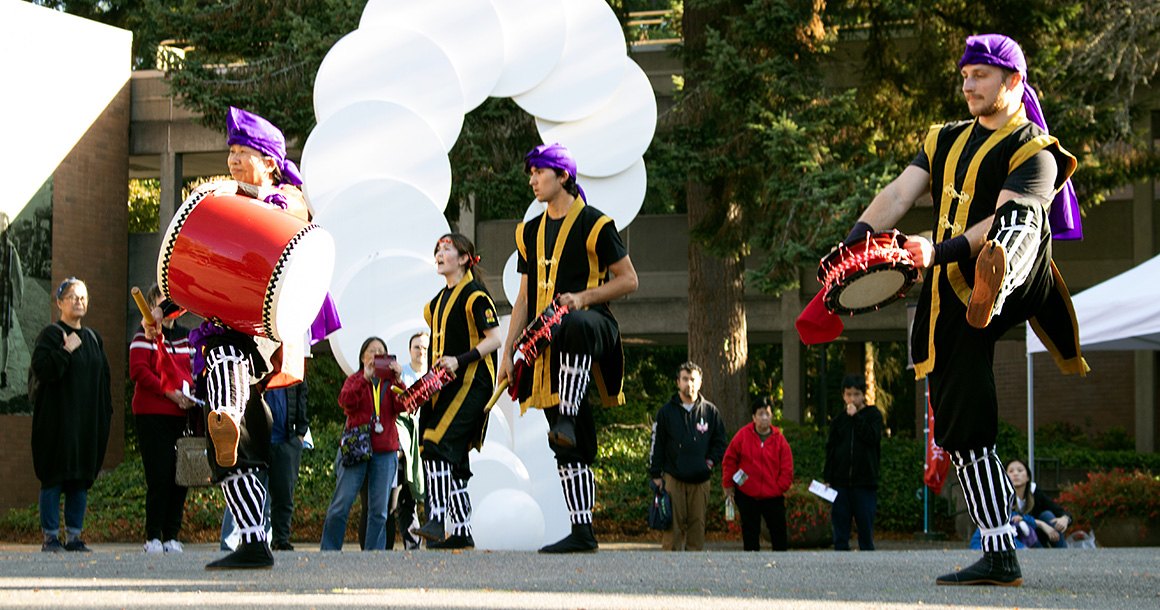 Performers in black-and-gold happi coats playing taiko drums outdoors before a crowd.