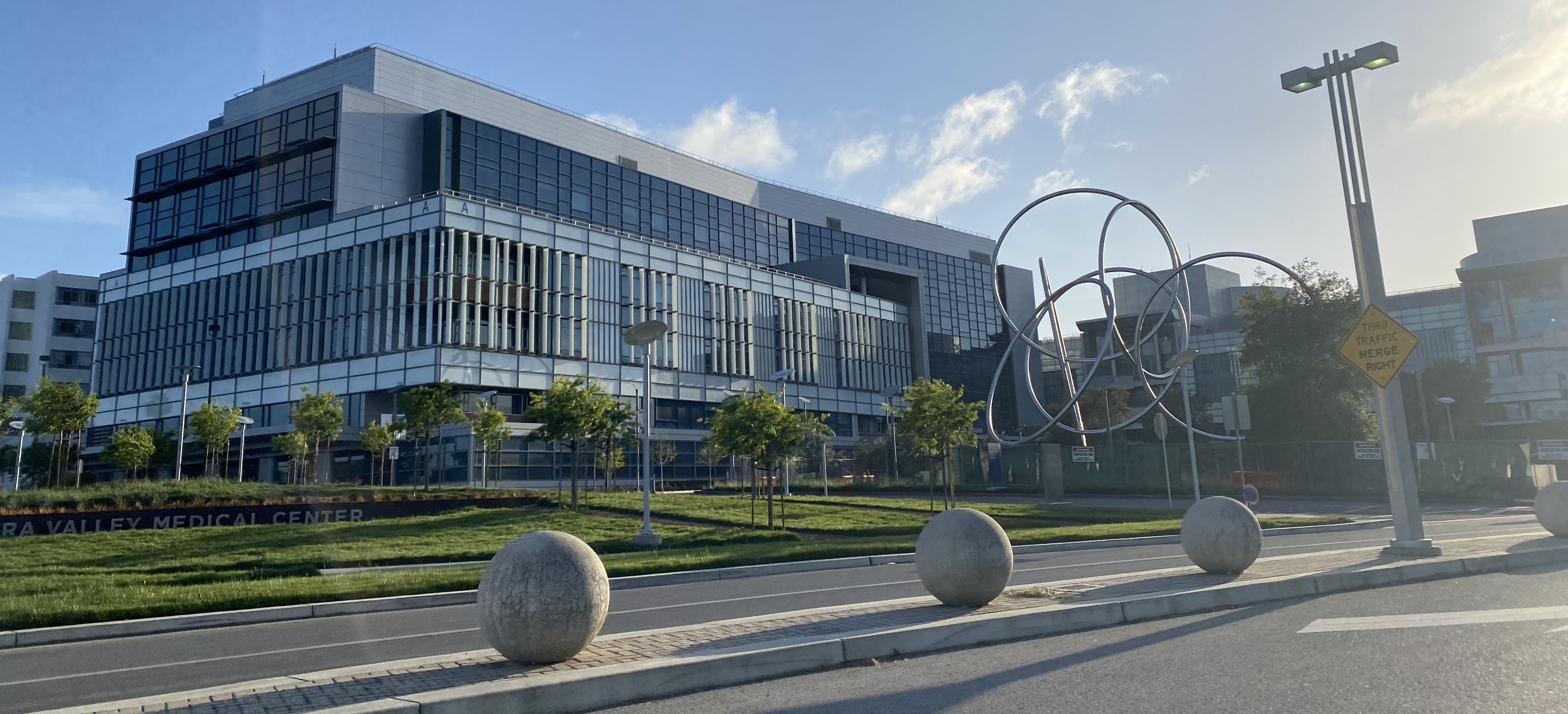 Modern multi-story hospital building with glass facade and large abstract metal sculpture on landscaped grounds.