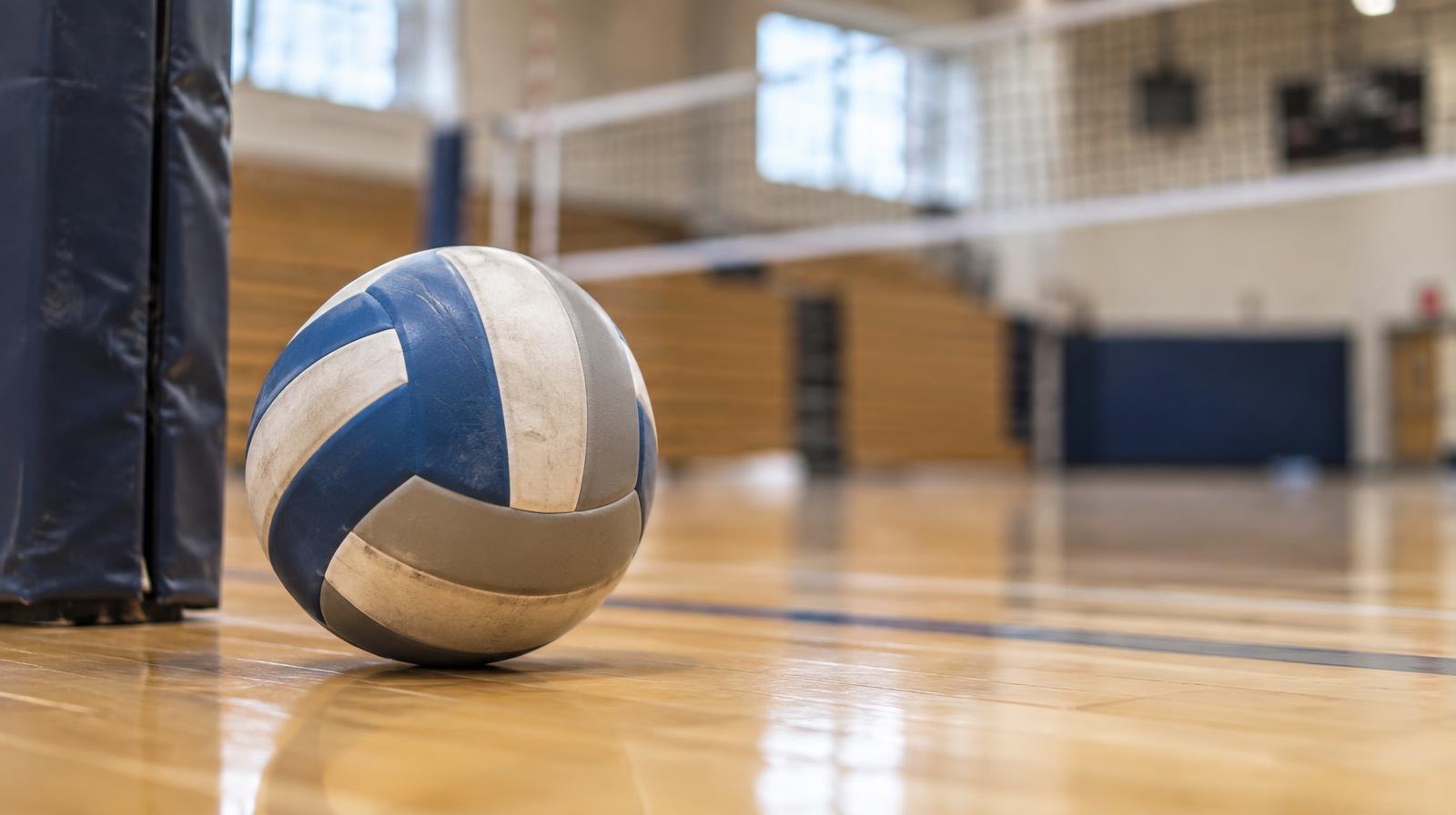 A volleyball resting on a hardwood gym floor near a net post.
