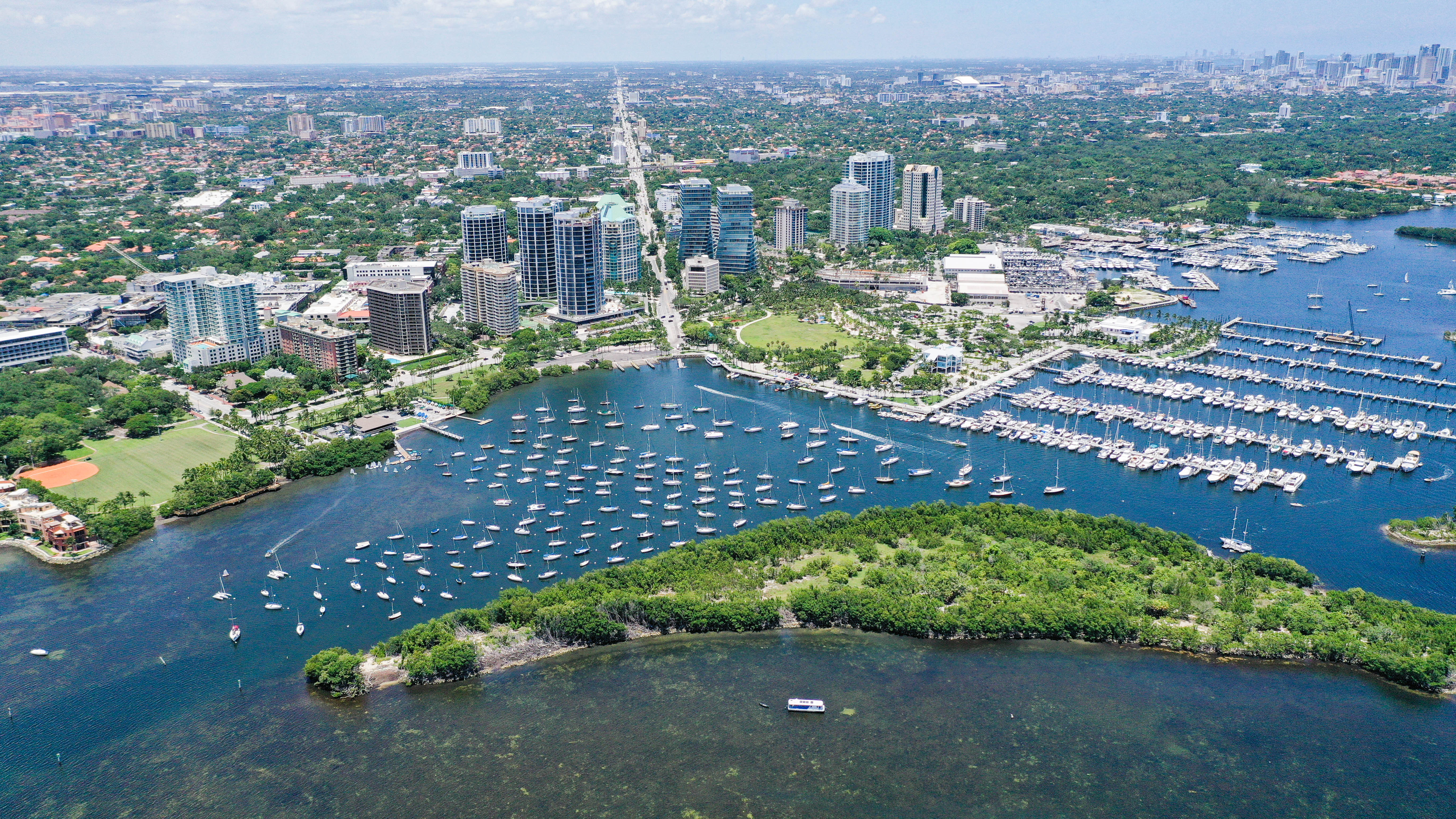 Aerial view of a marina filled with moored boats along a Miami waterfront with a city skyline beyond.