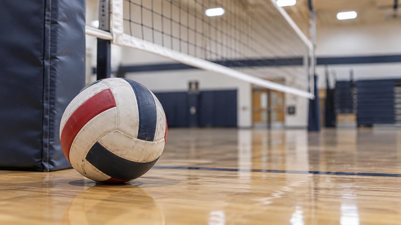 A volleyball sitting on a hardwood gym floor near a volleyball net.