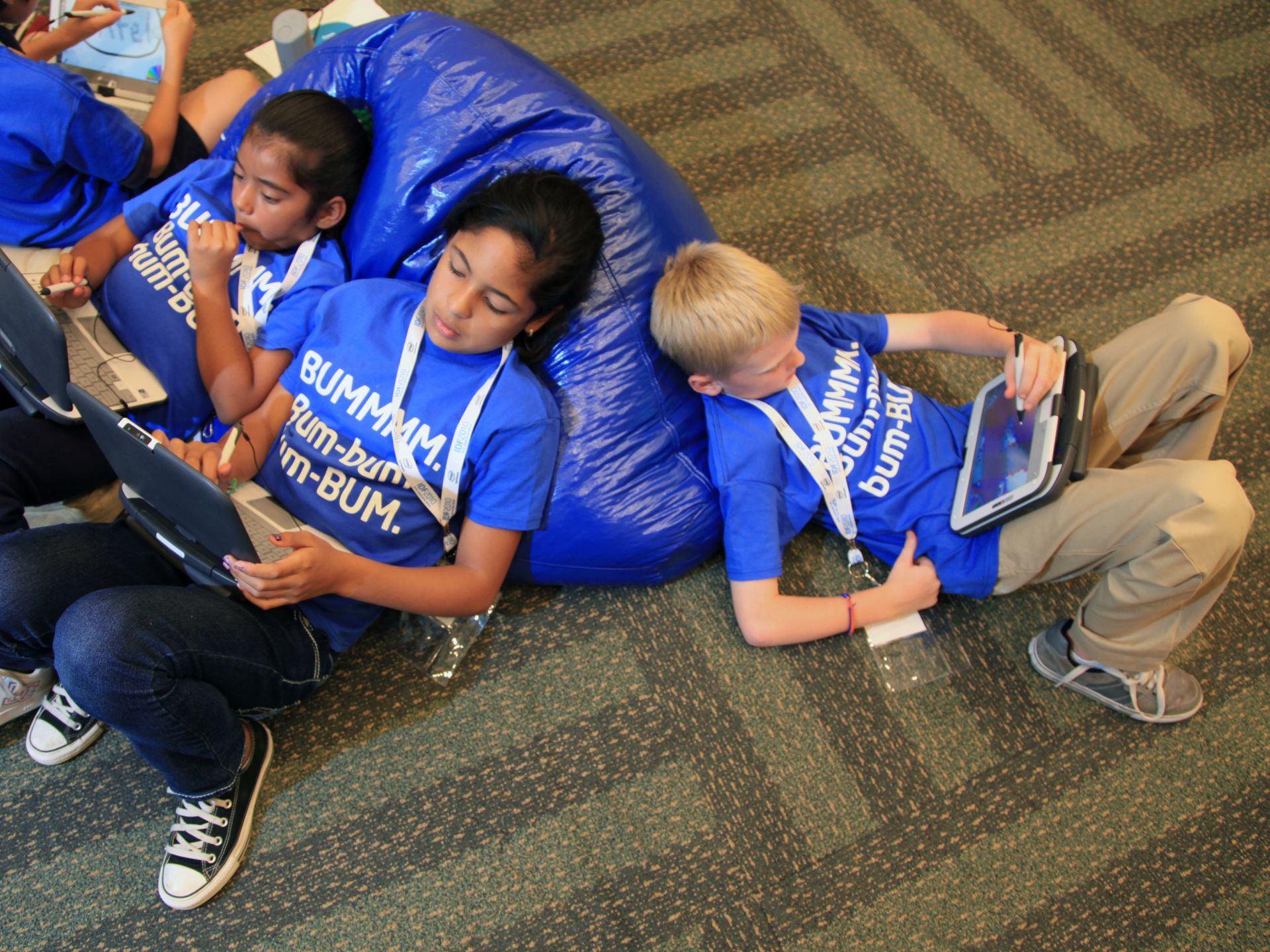 Three children in blue shirts use tablets and a laptop on a carpeted floor.