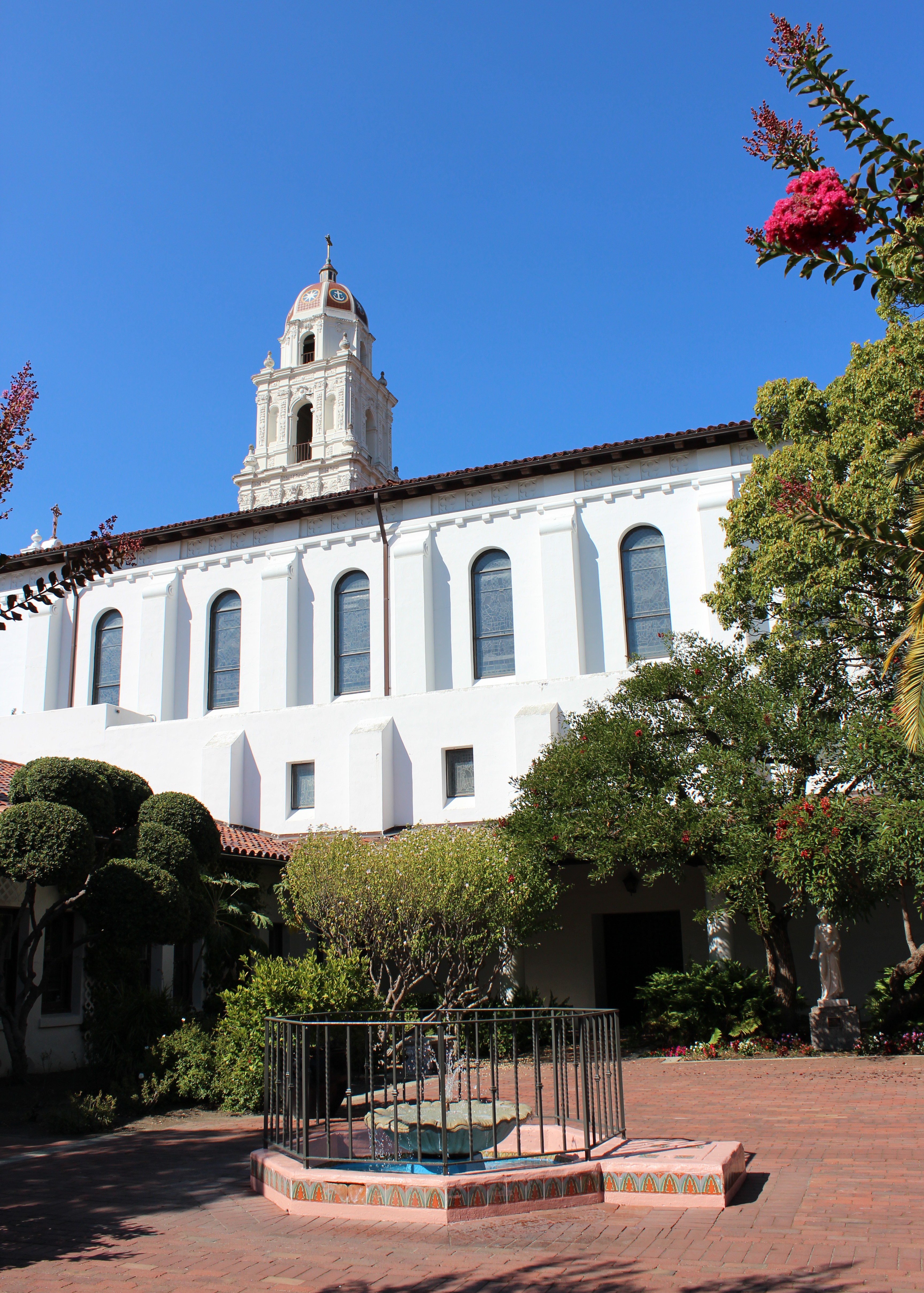 White Spanish Colonial Revival building with bell tower and courtyard fountain on a sunny day.