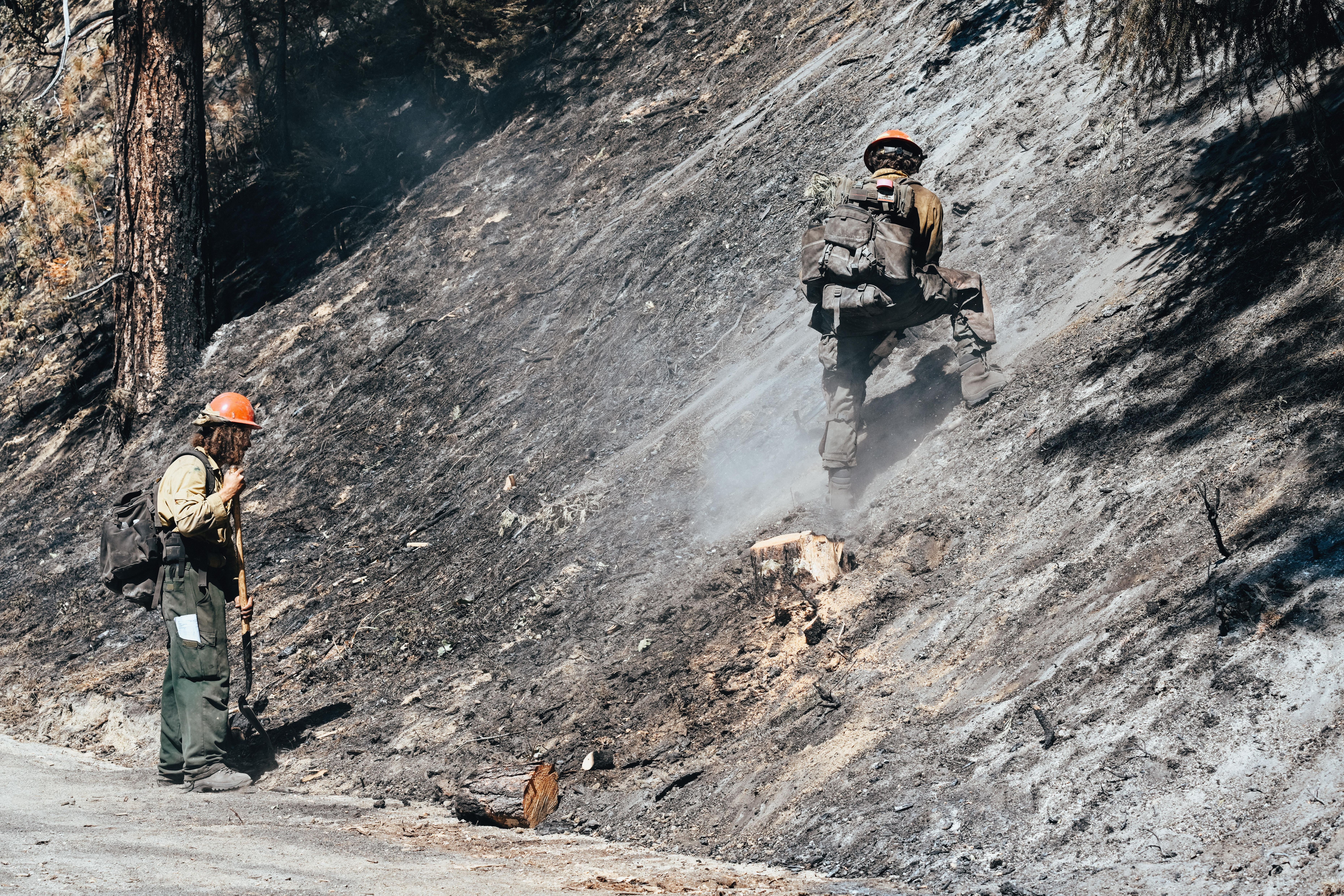 Two firefighters in hard hats work on a steep, smoke-covered burned hillside.
