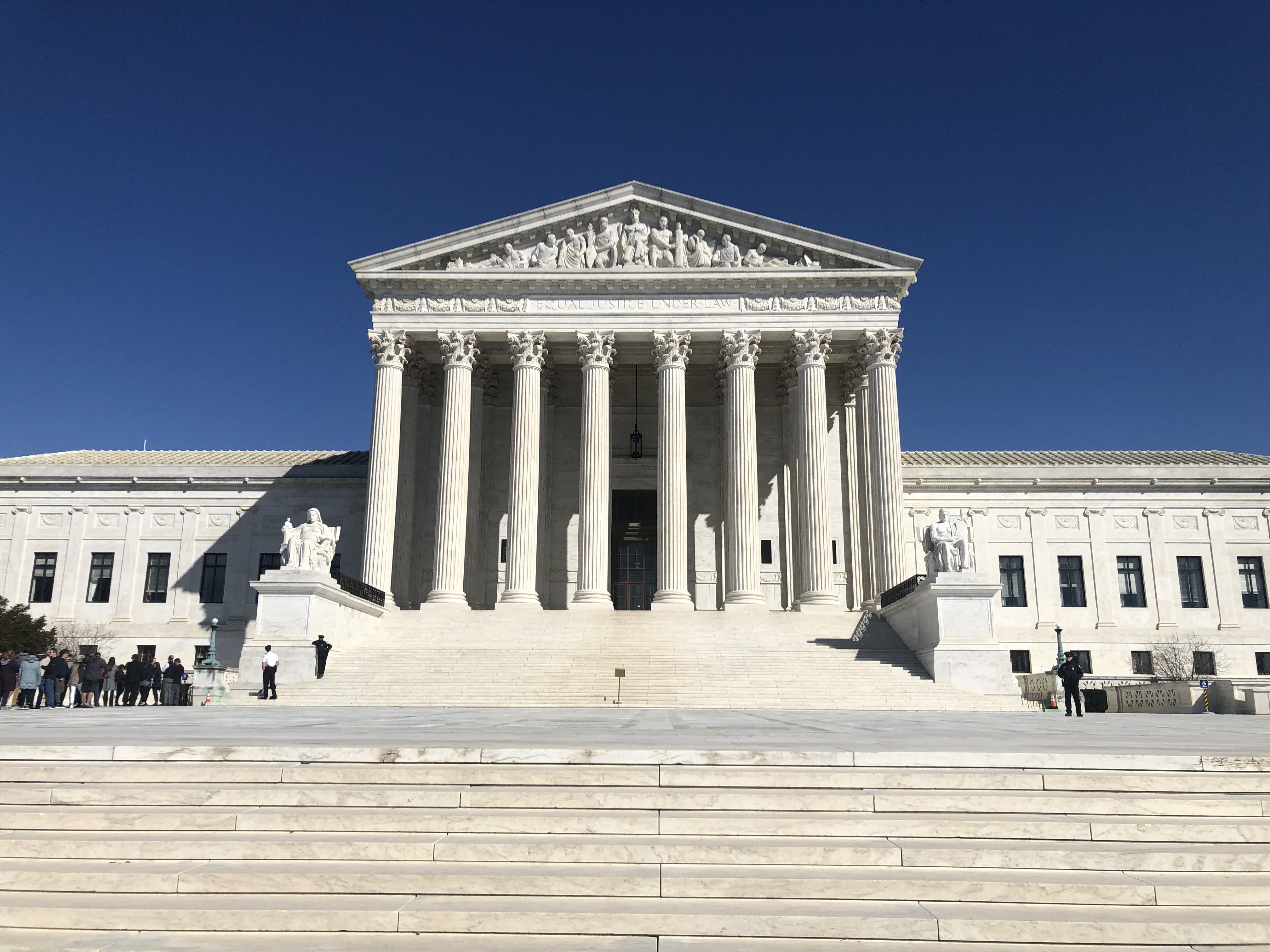 Front facade of the U.S. Supreme Court Building with tall columns and marble steps under a blue sky.