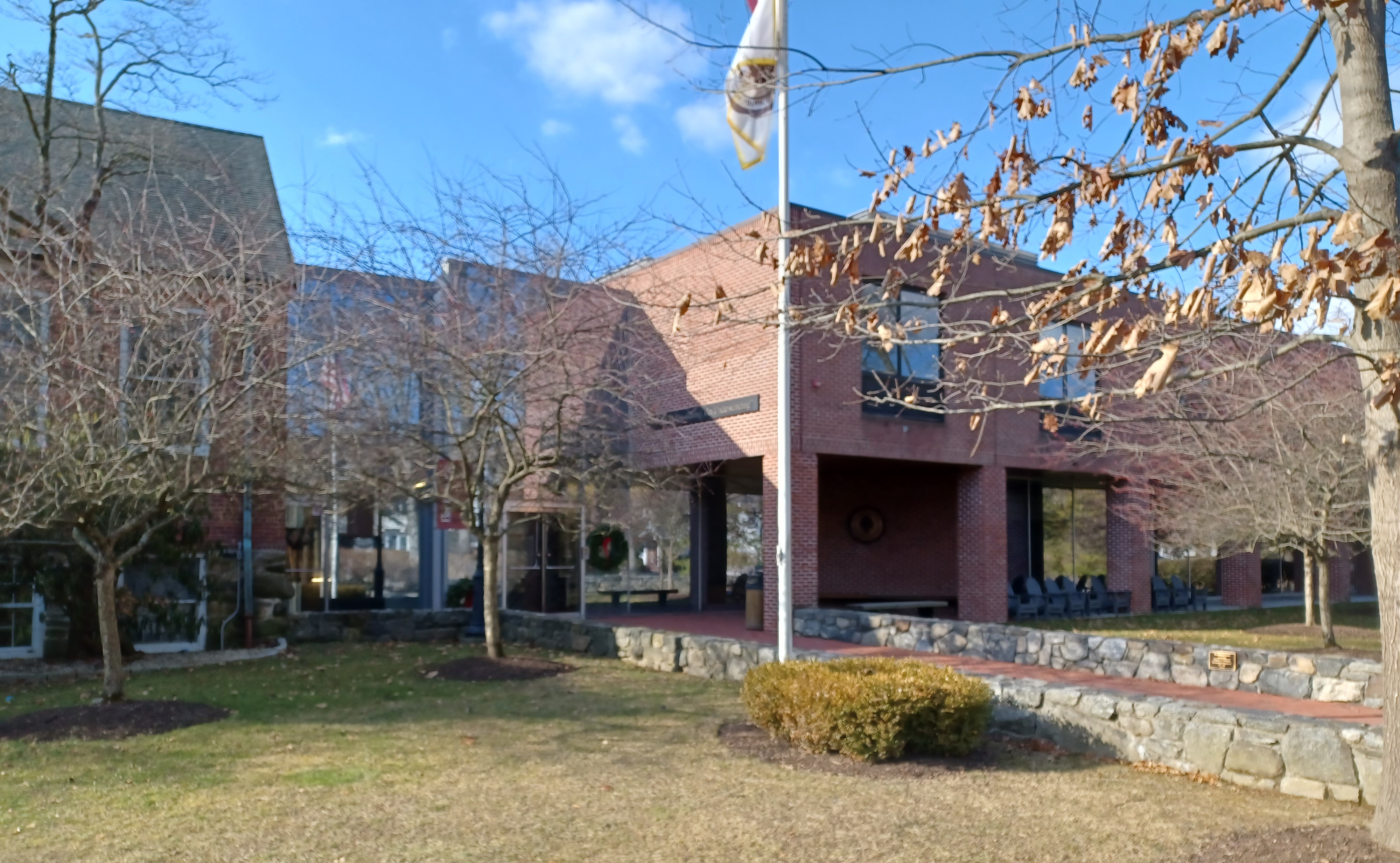 Brick school building with large glass windows, flagpole, and bare trees on a lawn.