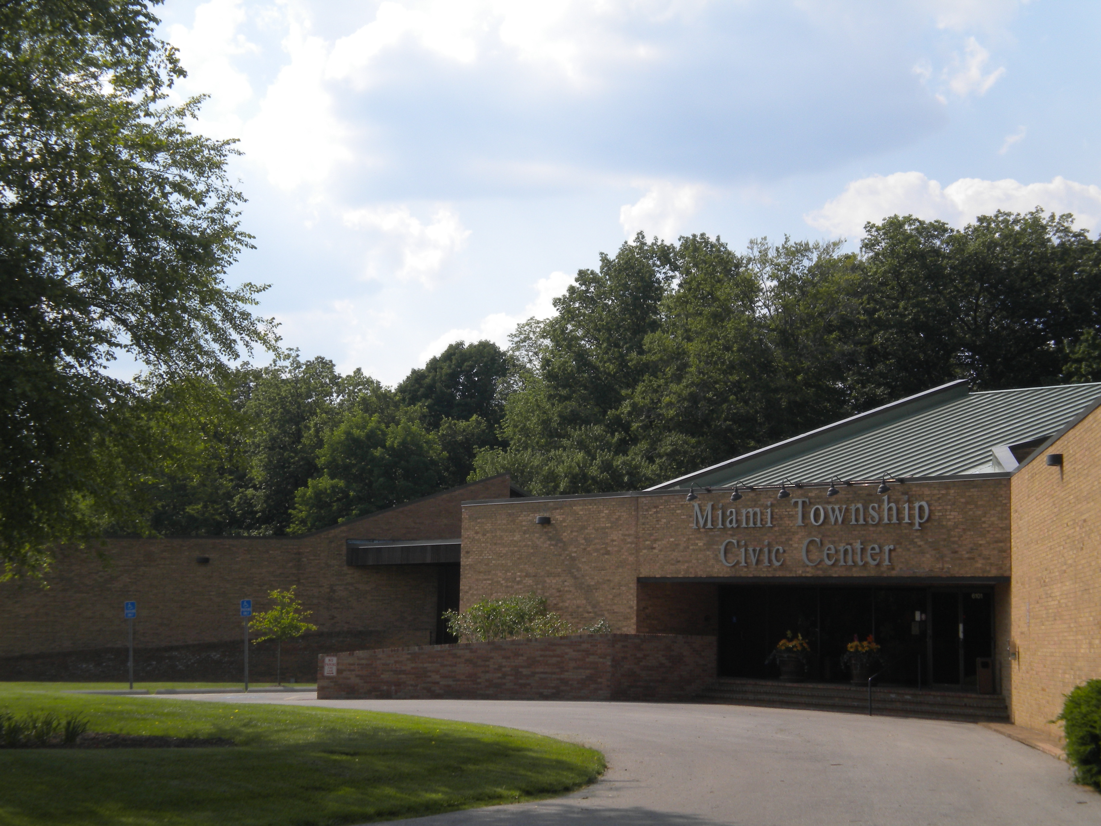 Brick building with green metal roof and curved driveway, surrounded by trees.