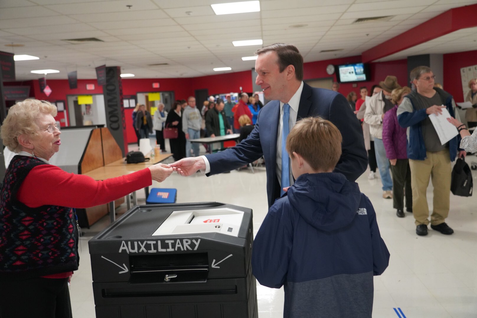 A man in a suit hands a document to an elderly poll worker at a voting station inside a polling place.