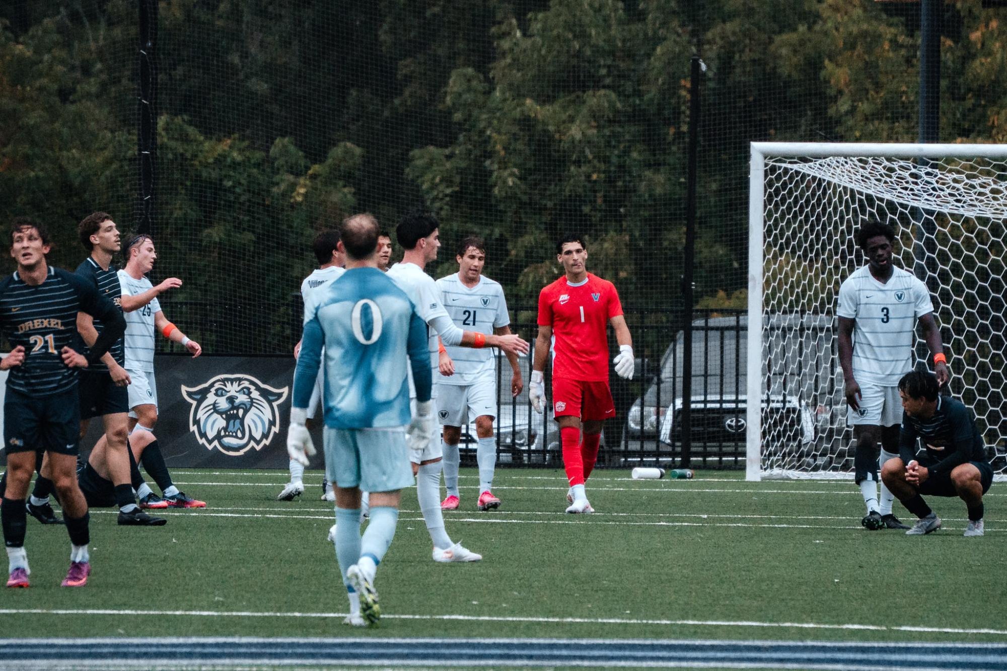 Soccer players in white and dark uniforms crowd near a goal on a turf field.