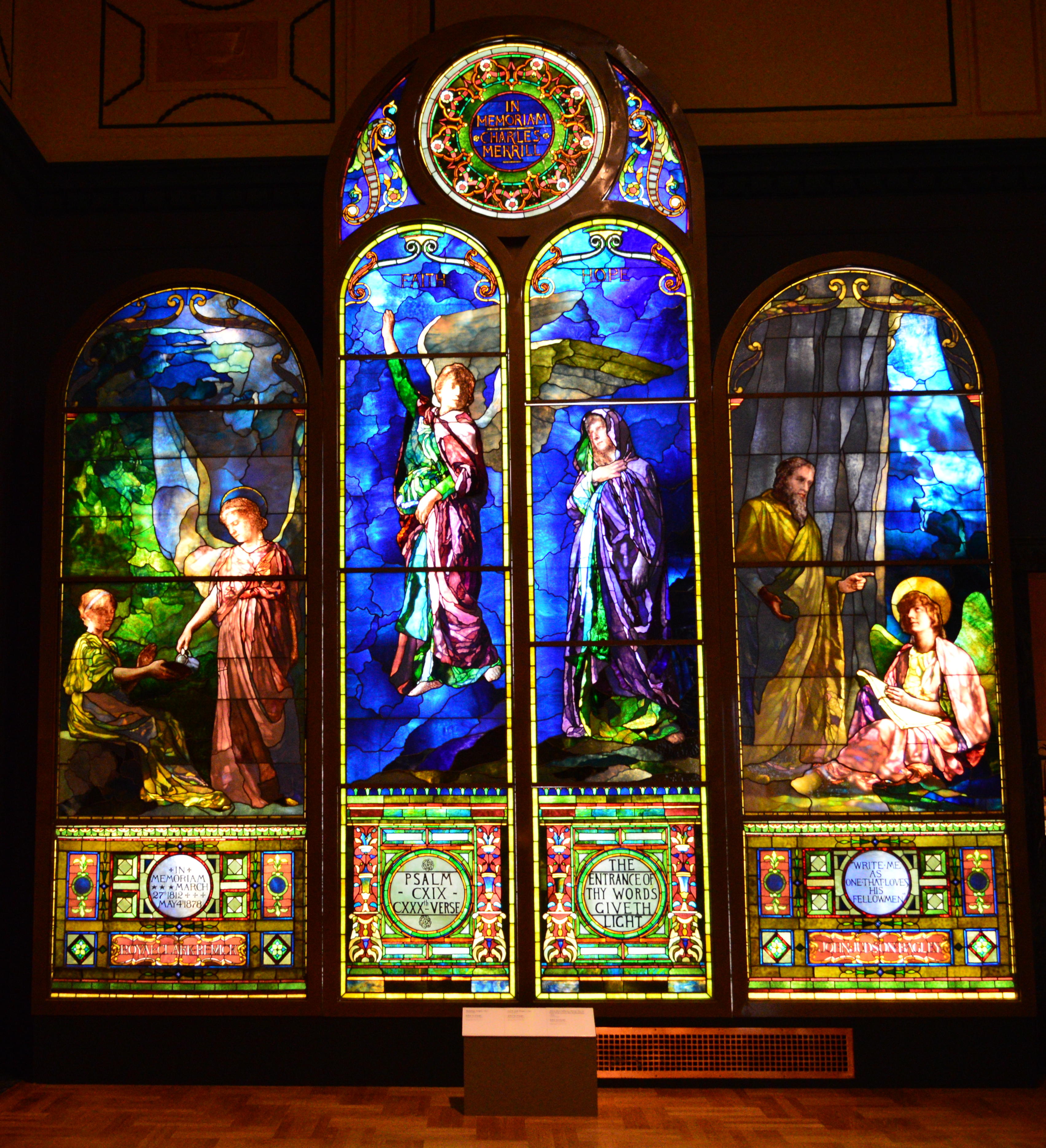 Four large arched stained glass windows with angelic figures displayed in a darkened museum gallery.