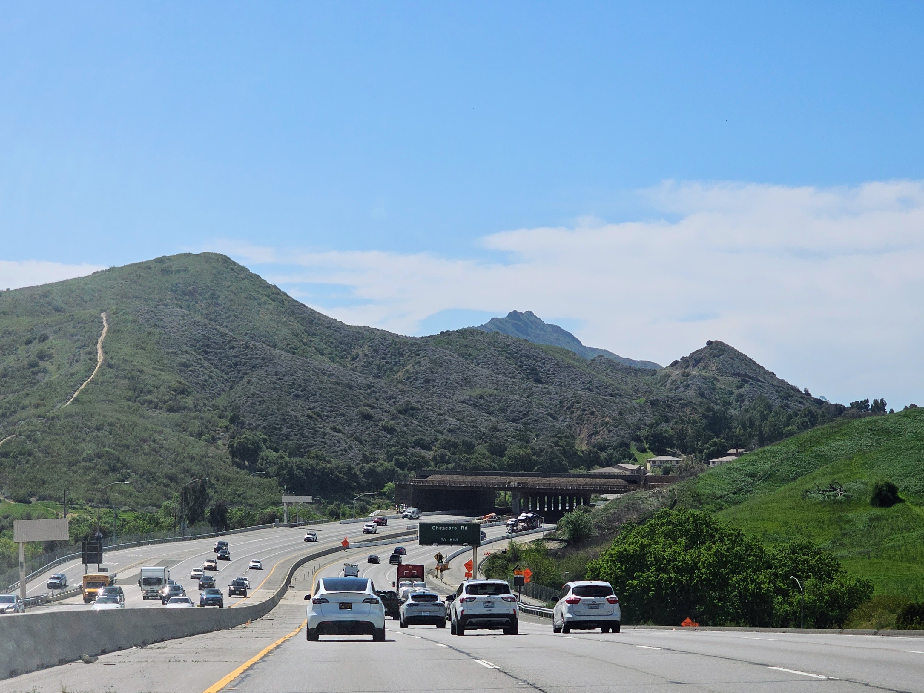 Multi-lane freeway with a wide overpass bridge spanning it, green hills and mountain in background.