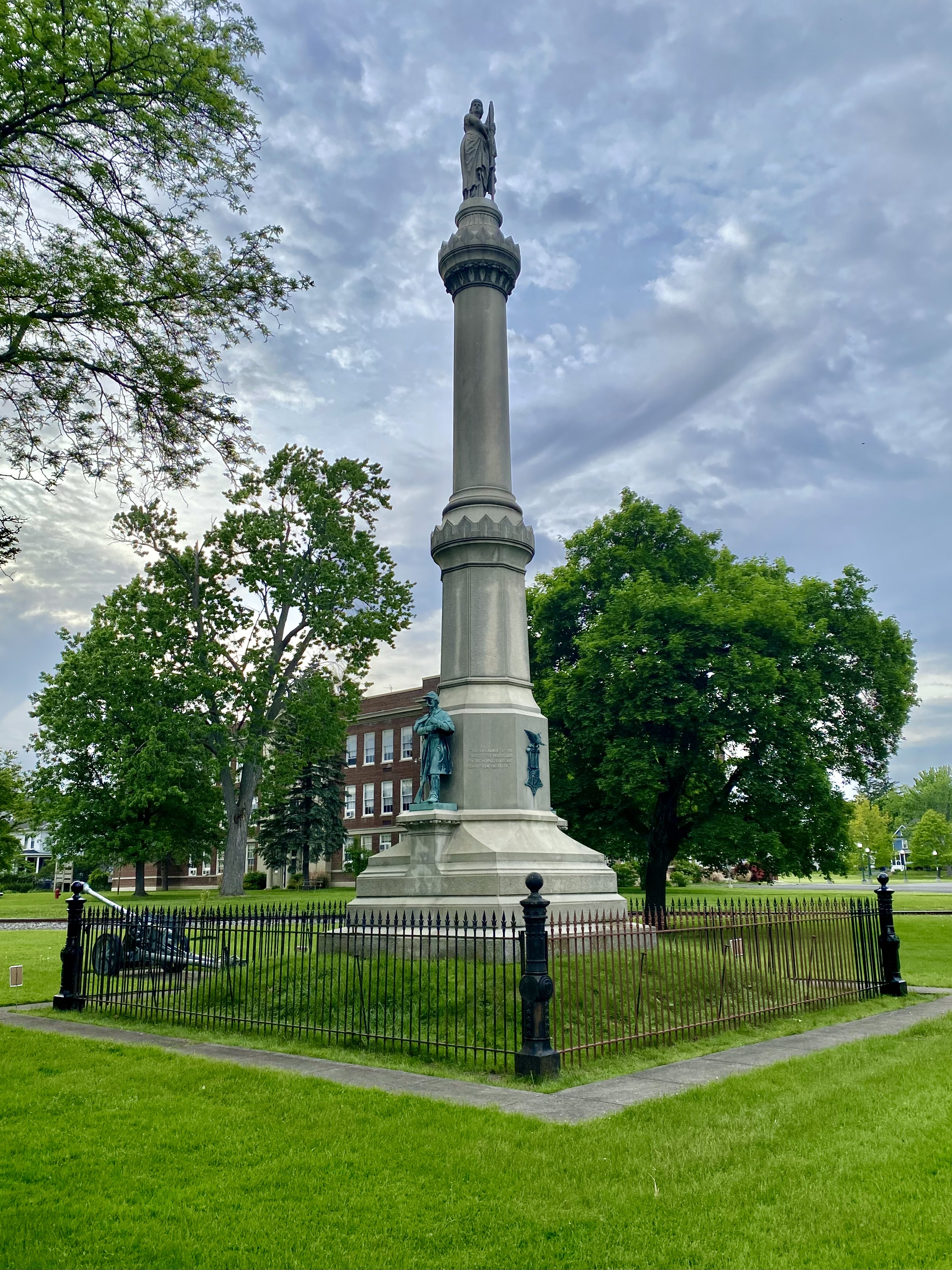 Tall granite memorial column with bronze figures stands inside an iron fence in a park.