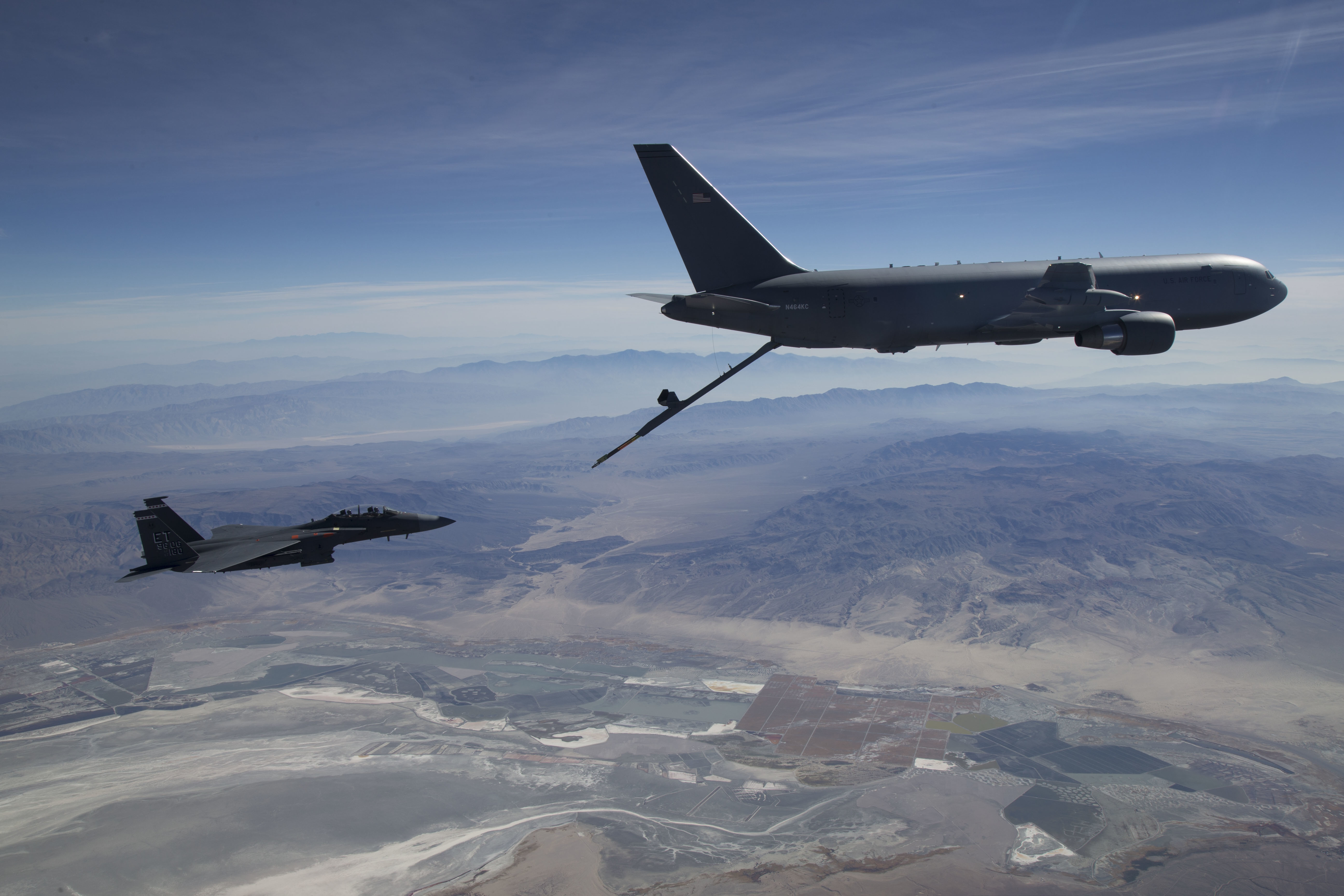 A military tanker aircraft extends a refueling boom toward a fighter jet in flight.