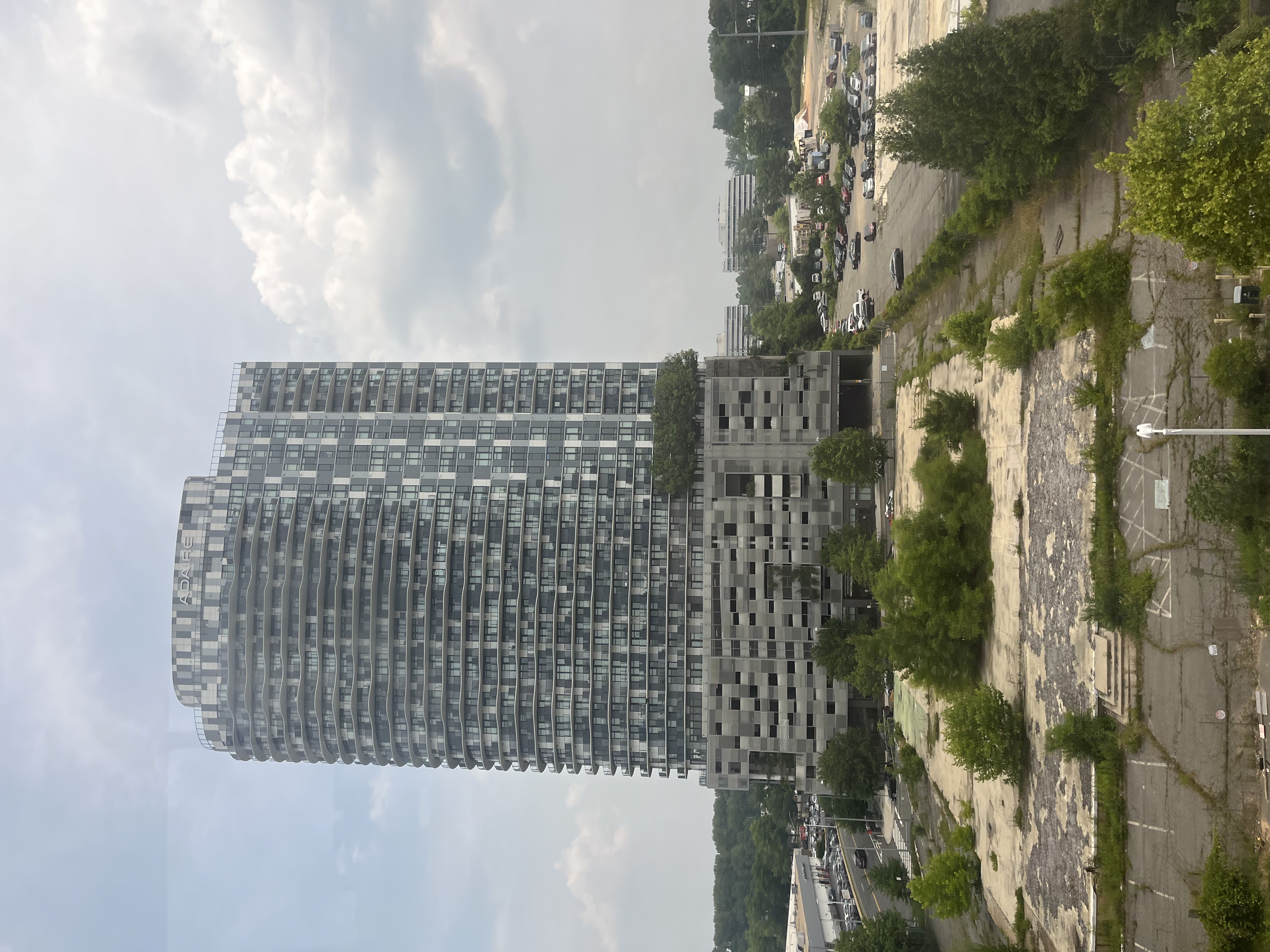 A tall modern high-rise apartment building surrounded by undeveloped land and lower-rise buildings under a partly cloudy sky.