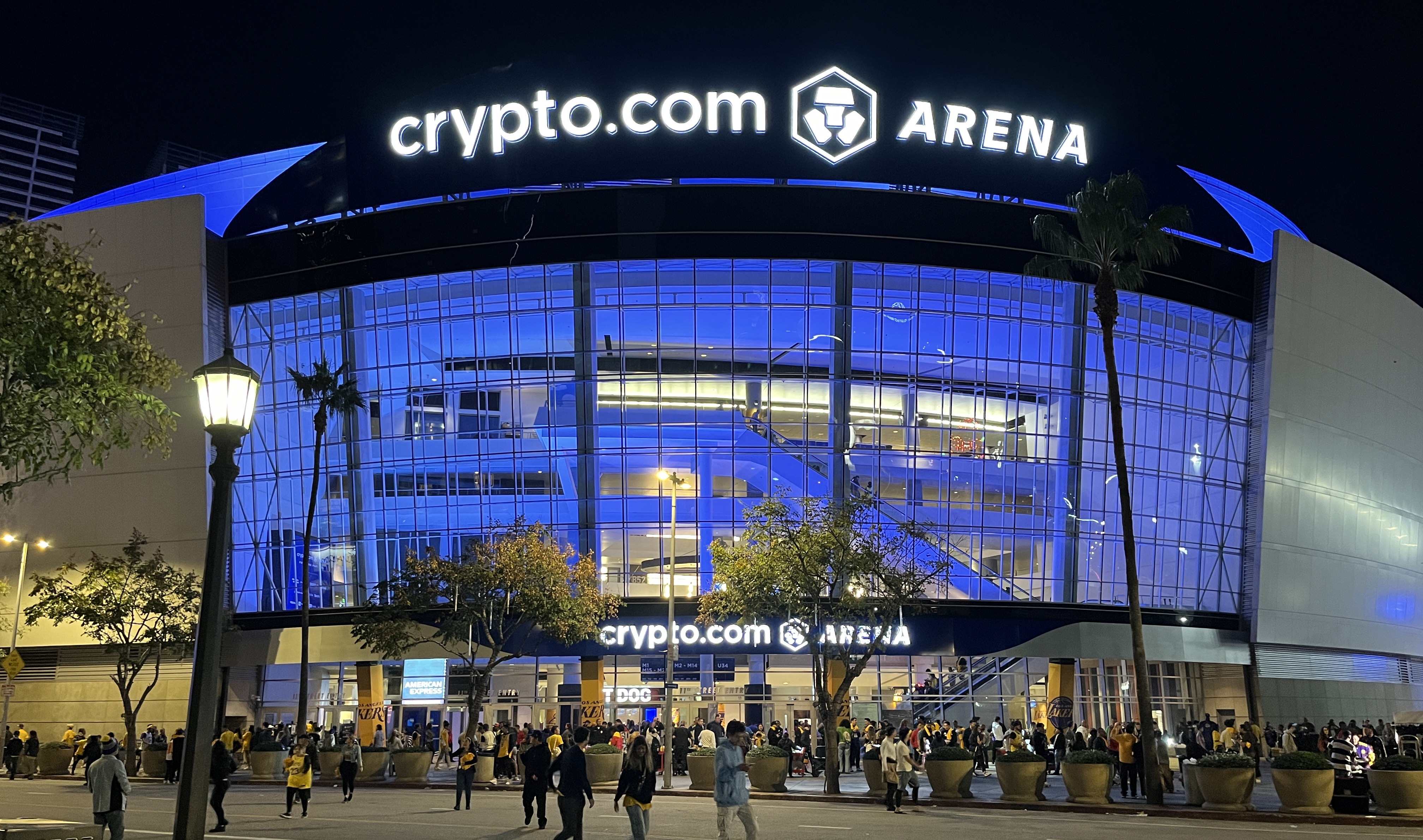 Arena exterior lit in blue at night with crowds and palm trees outside.