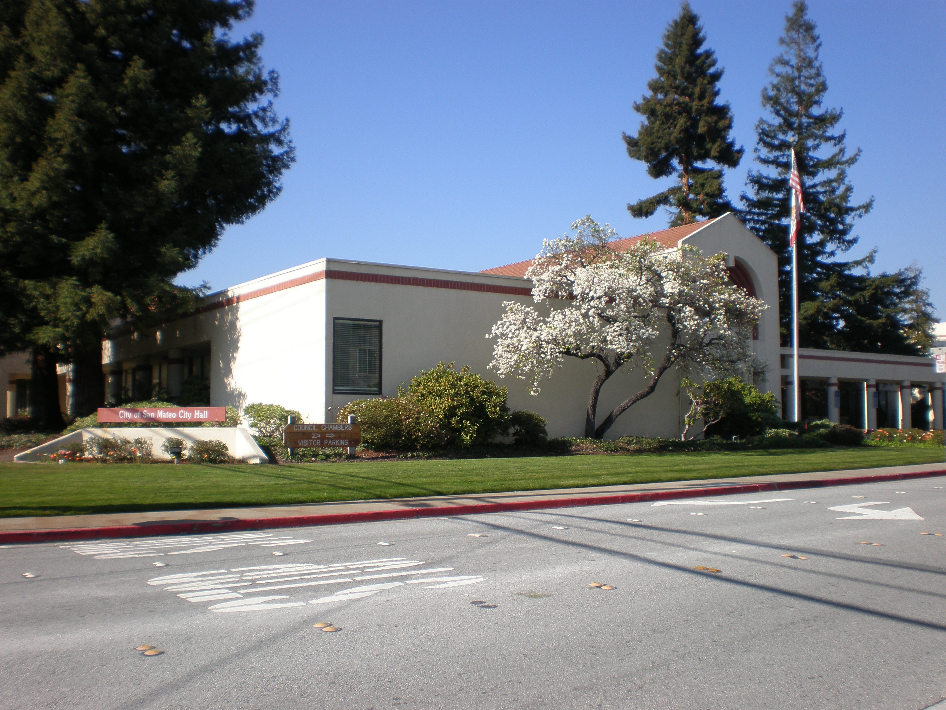 Exterior of a low-rise stucco government building with a flowering white tree and American flag.