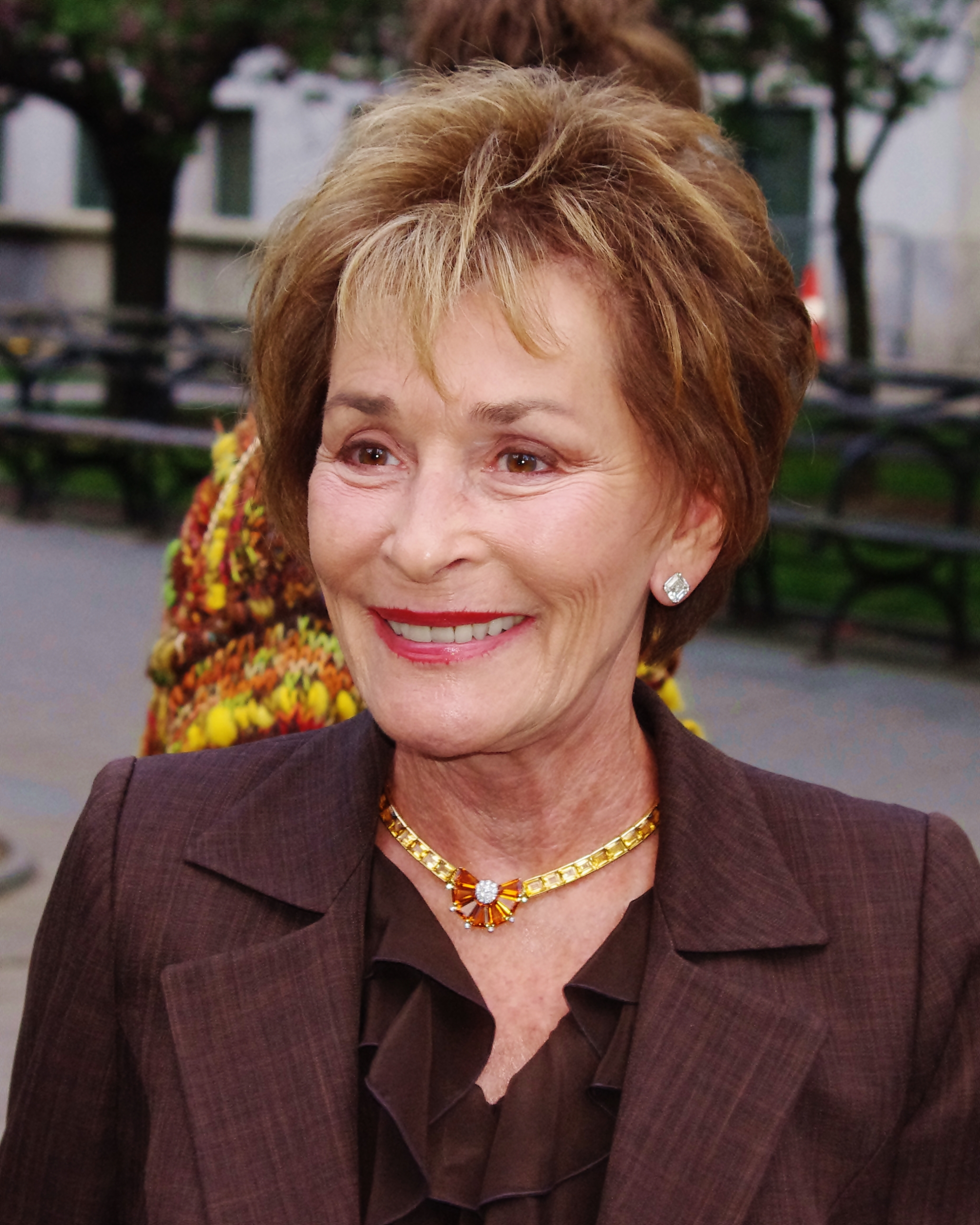 Woman in brown blazer and gold necklace smiling at an outdoor event.