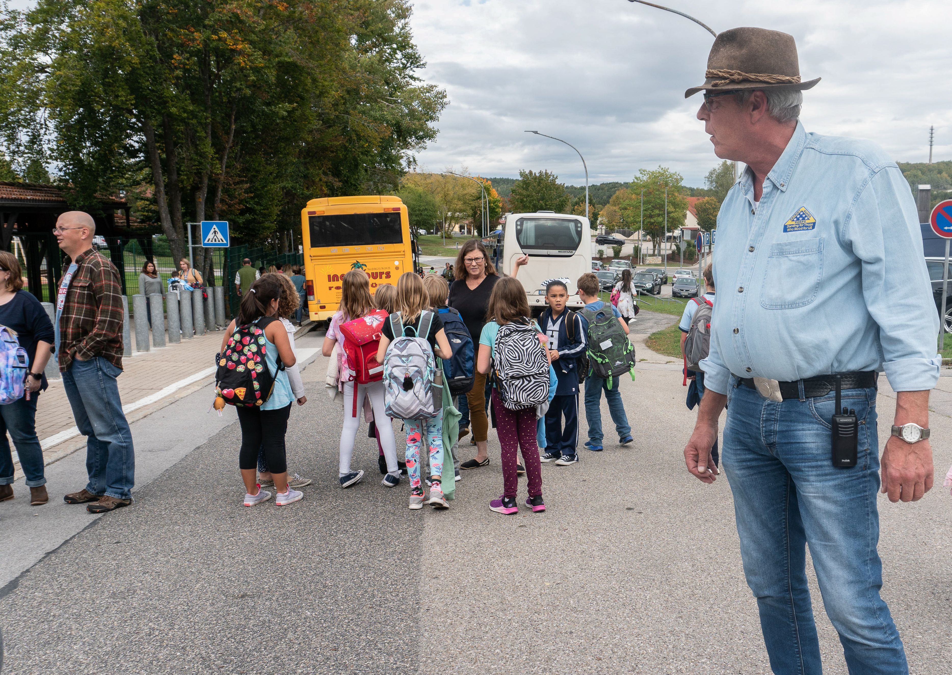 A man watches children with backpacks walk toward a yellow school bus.