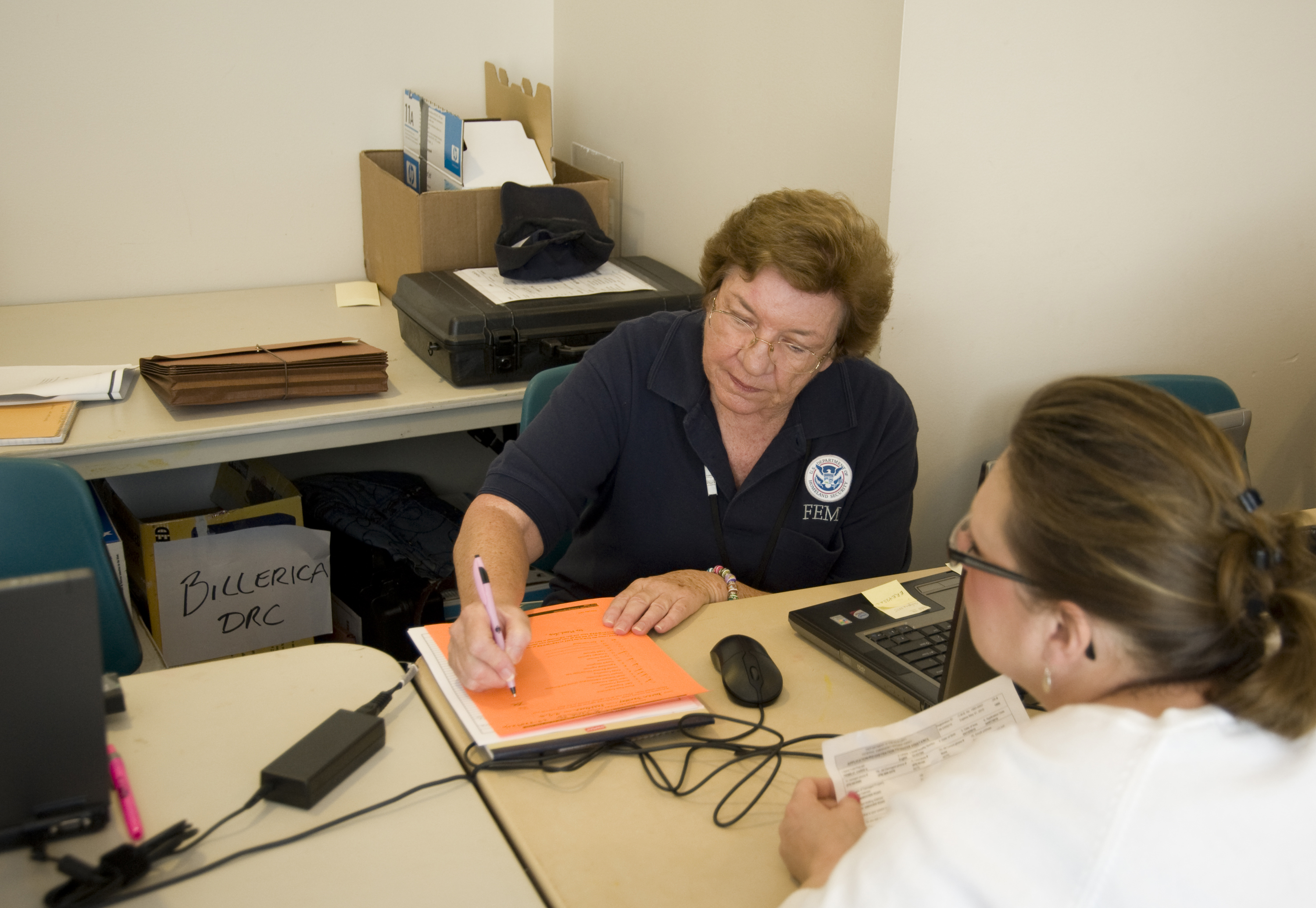 FEMA specialist in navy polo shirt writes on a notepad while helping an applicant at a desk.