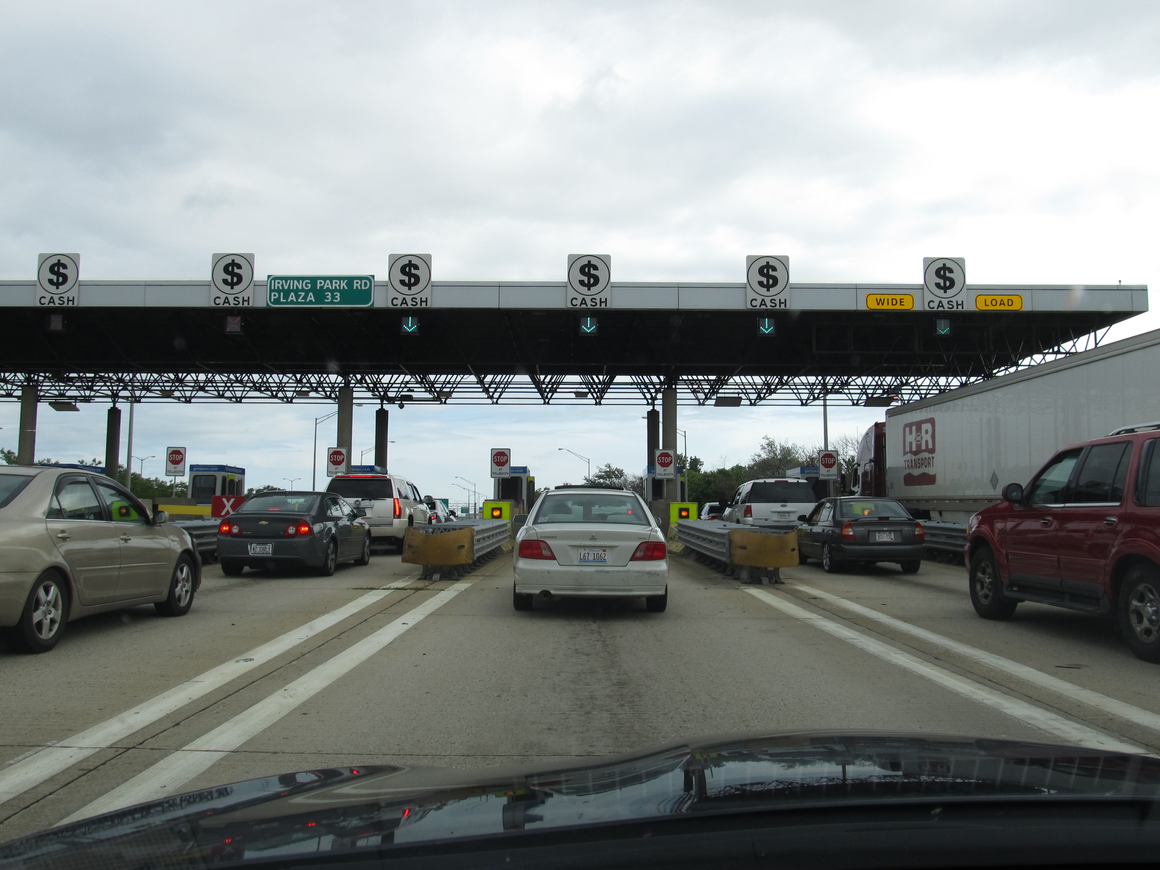 Cars and trucks lined up at a multi-lane cash toll plaza on an Illinois tollway.