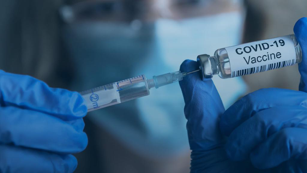 Gloved hands drawing vaccine from a labeled COVID-19 vial into a syringe.