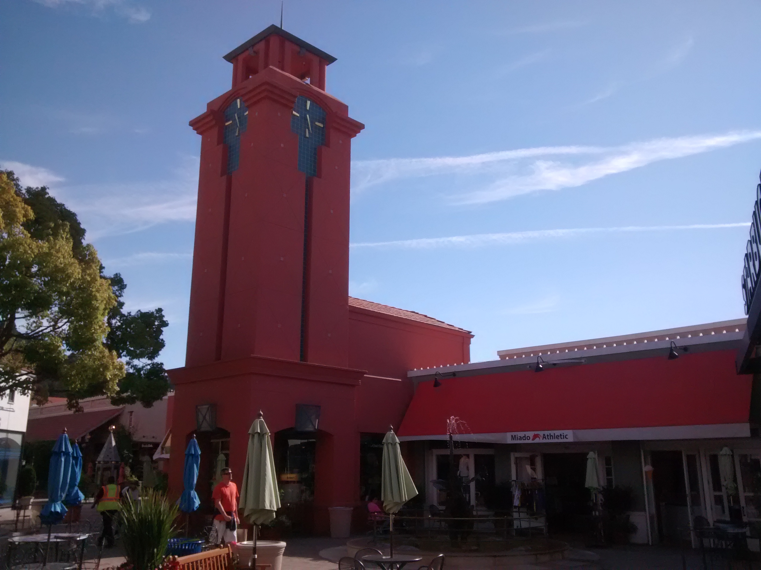 Red clock tower and retail storefronts at an outdoor shopping center.