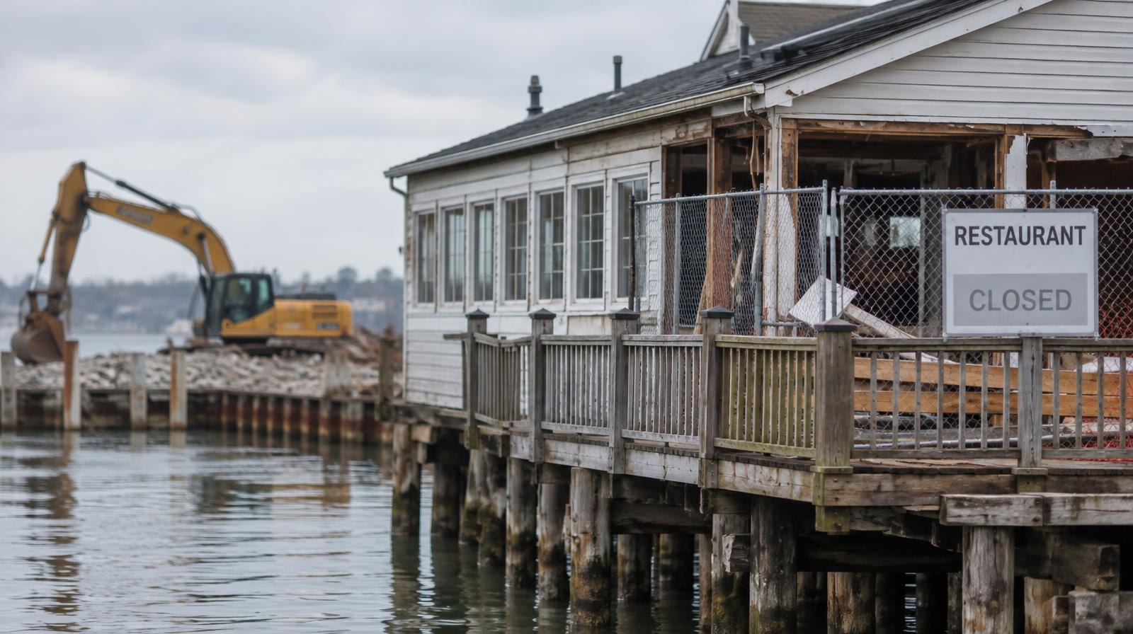 Weathered waterfront building on pilings with construction fencing nearby.