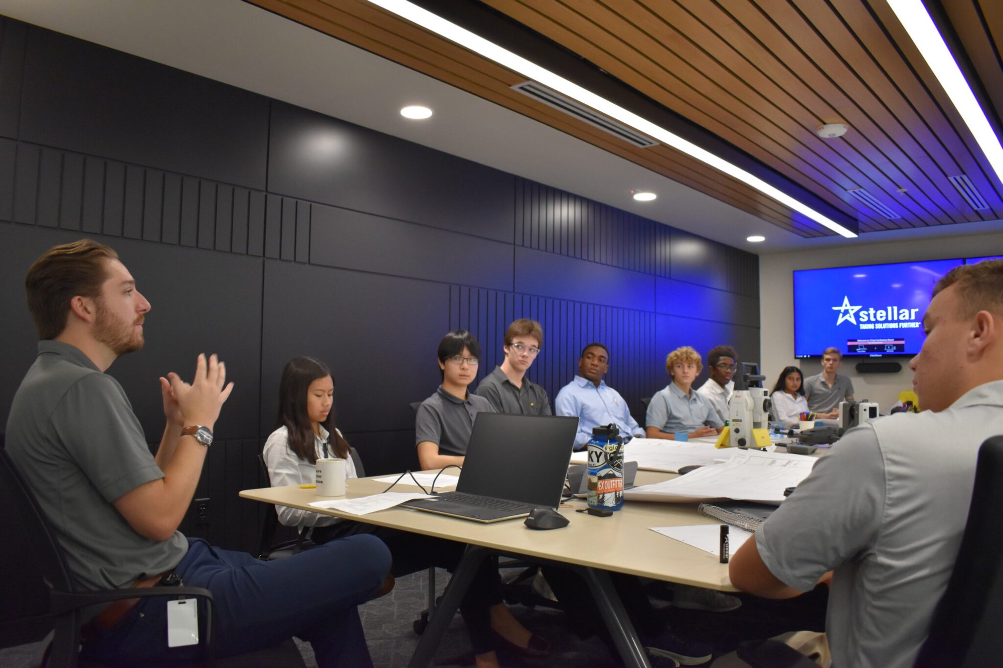 Students seated around a conference table in a modern meeting room with a company logo on a large screen.