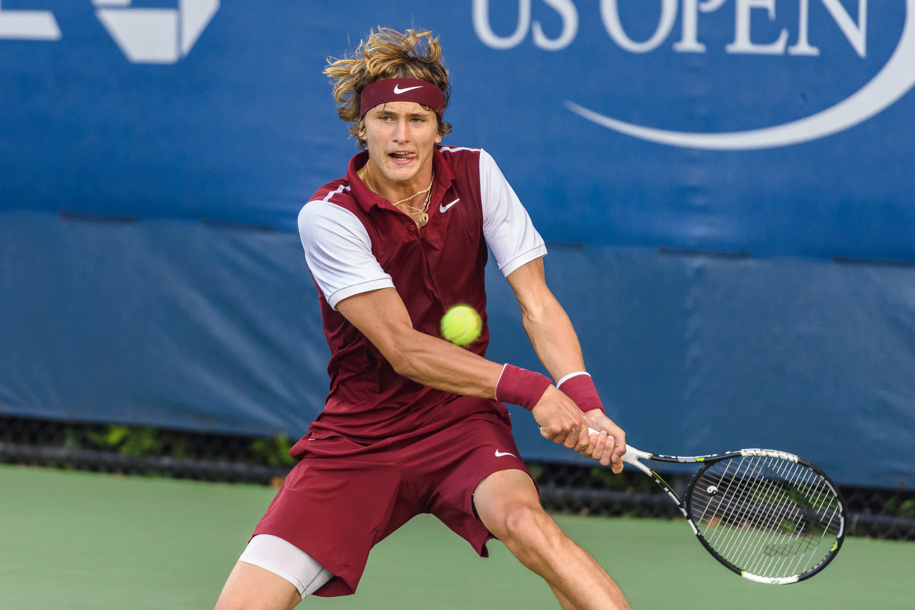 Male tennis player in maroon outfit hits a backhand with a US Open banner behind him.