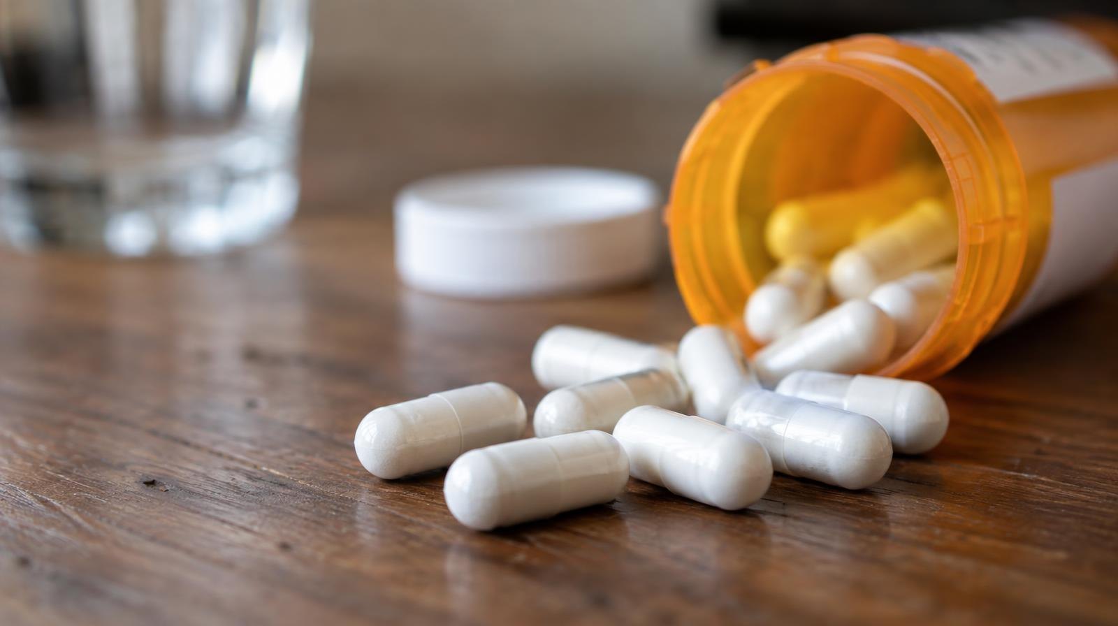 Orange prescription pill bottle tipped over with white capsules spilling onto a wooden surface.
