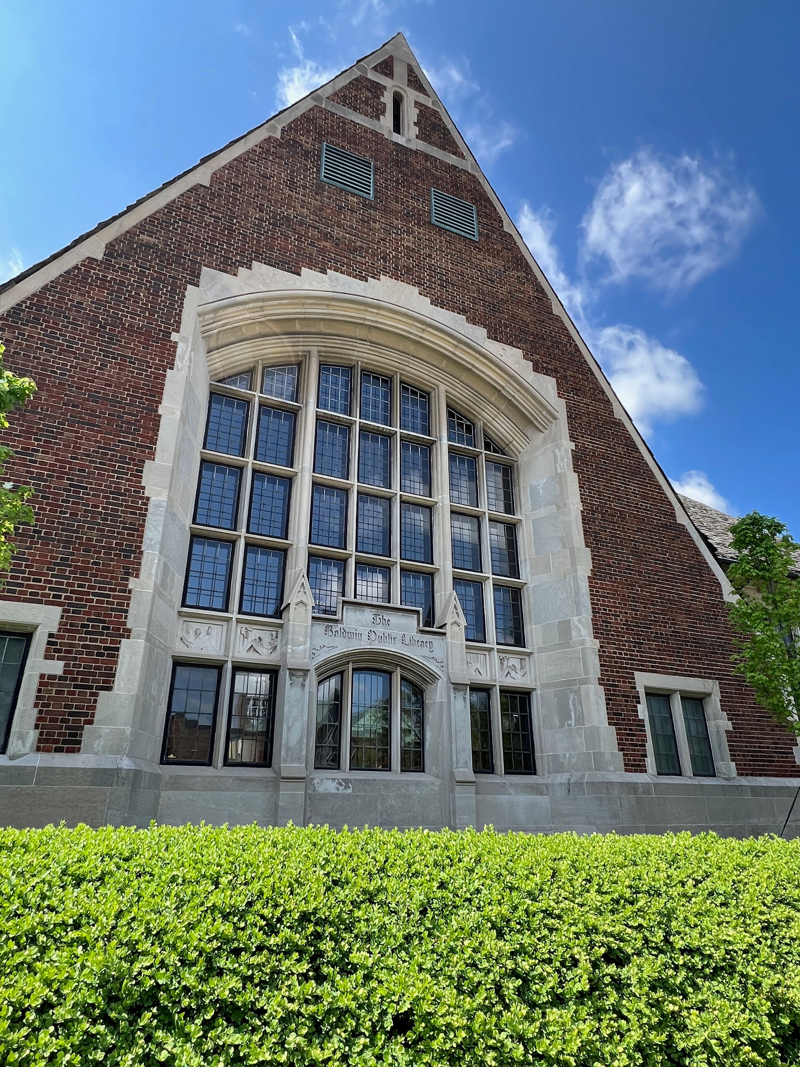 Red-brick Gothic library building with arched window and trimmed hedges in front.