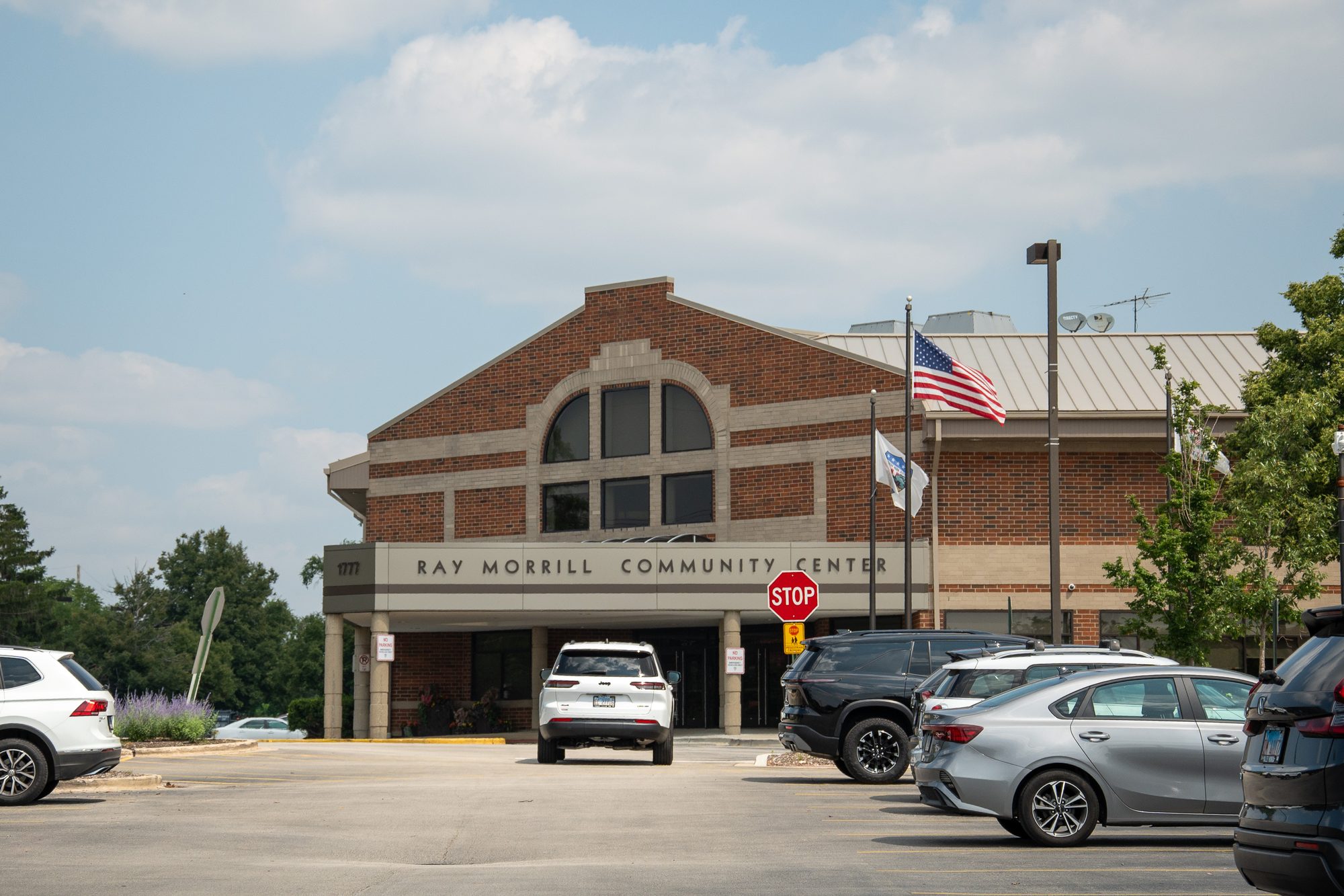 Brick community center building with arched window, flags, and parking lot.
