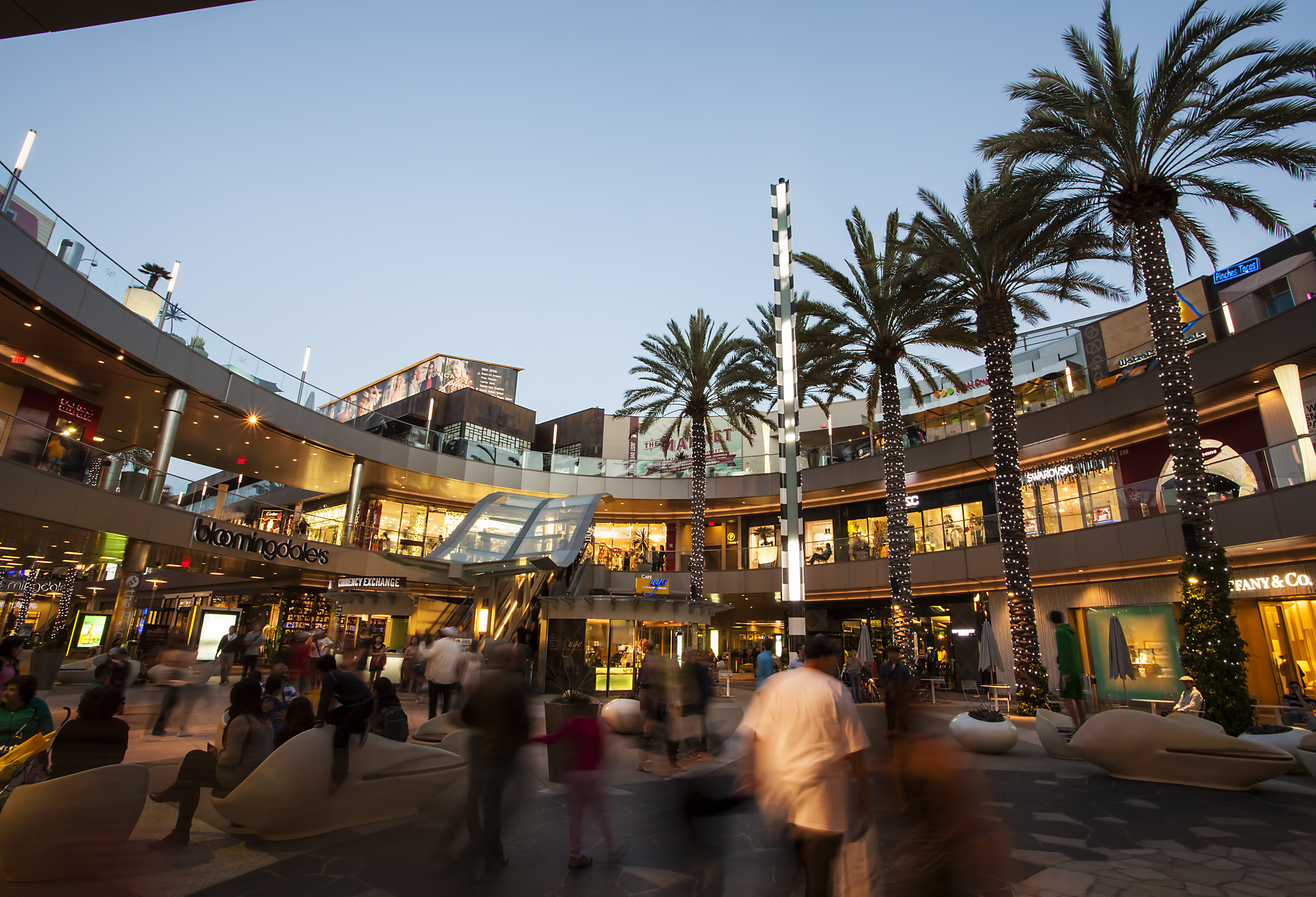 Multi-level open-air mall courtyard with palm trees and shoppers at dusk.