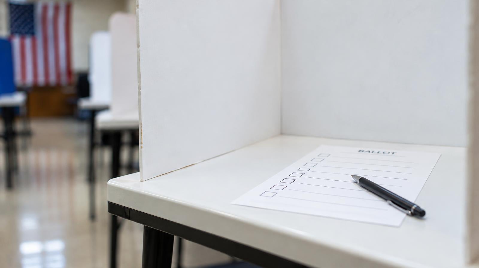 A blank paper ballot inside a white privacy voting booth with a pen.