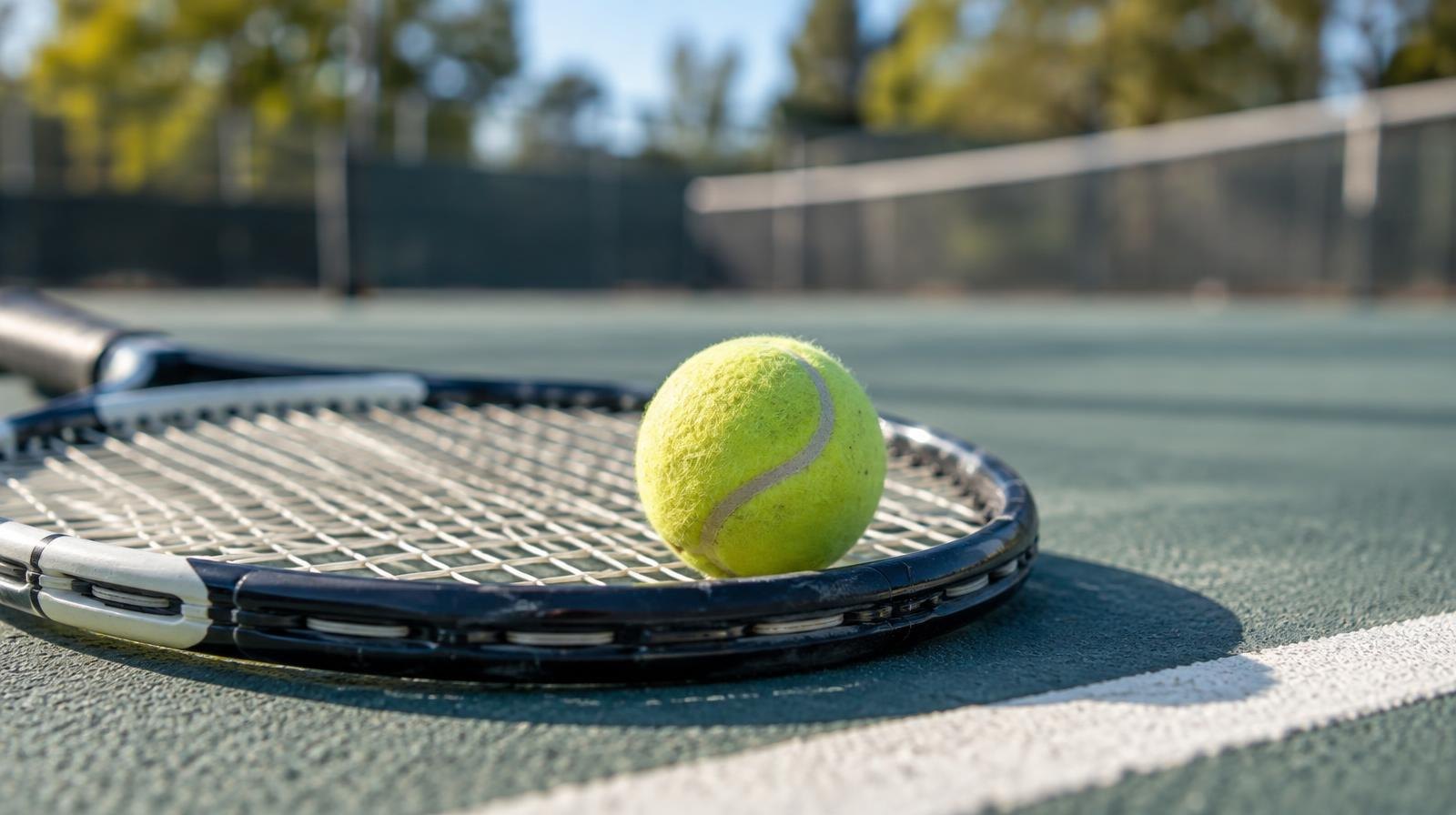 A yellow tennis ball resting on the strings of a tennis racket on a hard court surface.